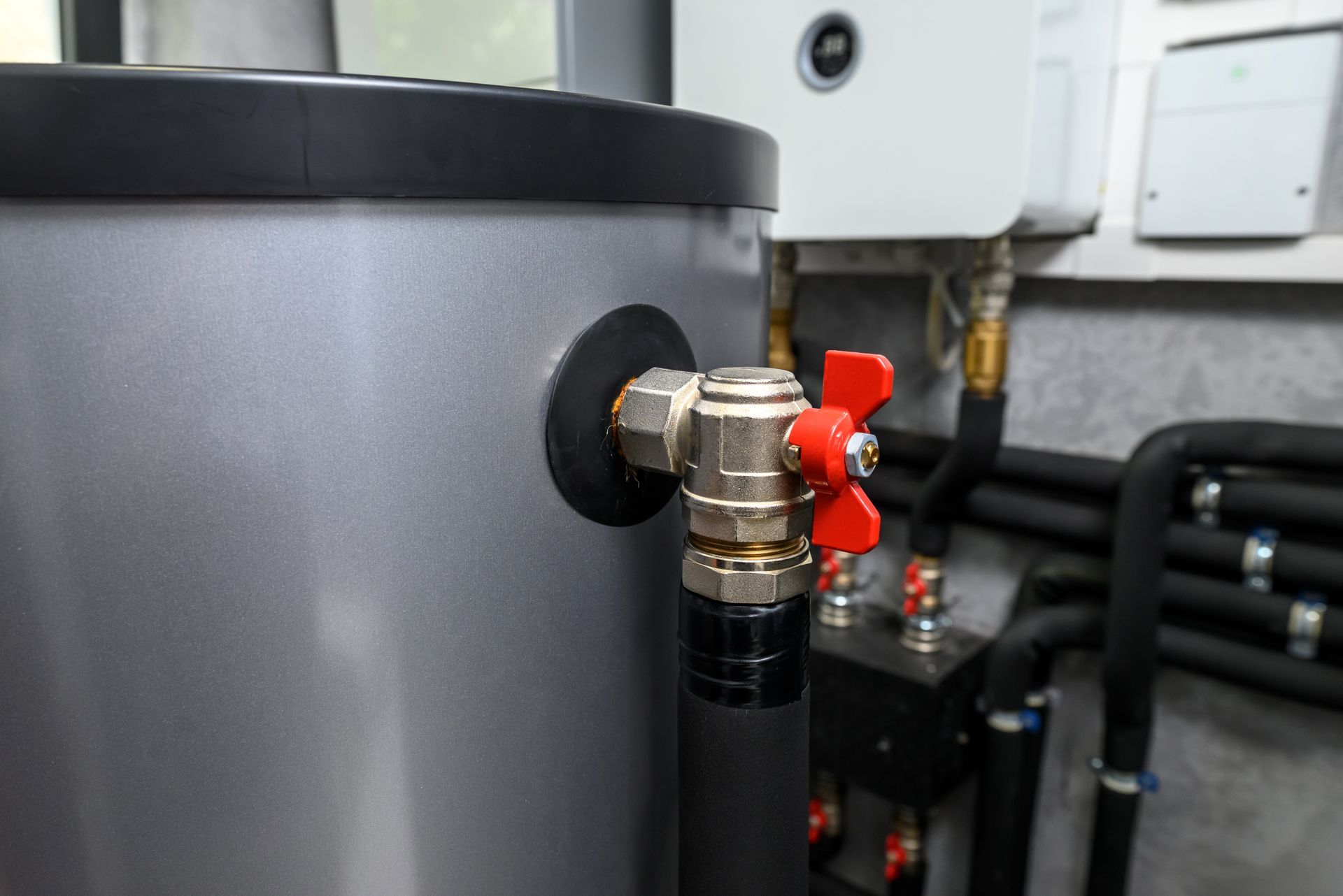 Close-up of a silver water tank with a red-handled valve and black pipe attached, indoors. Close-up of a silver water tank with a red-handled valve and black pipe attached, indoors.