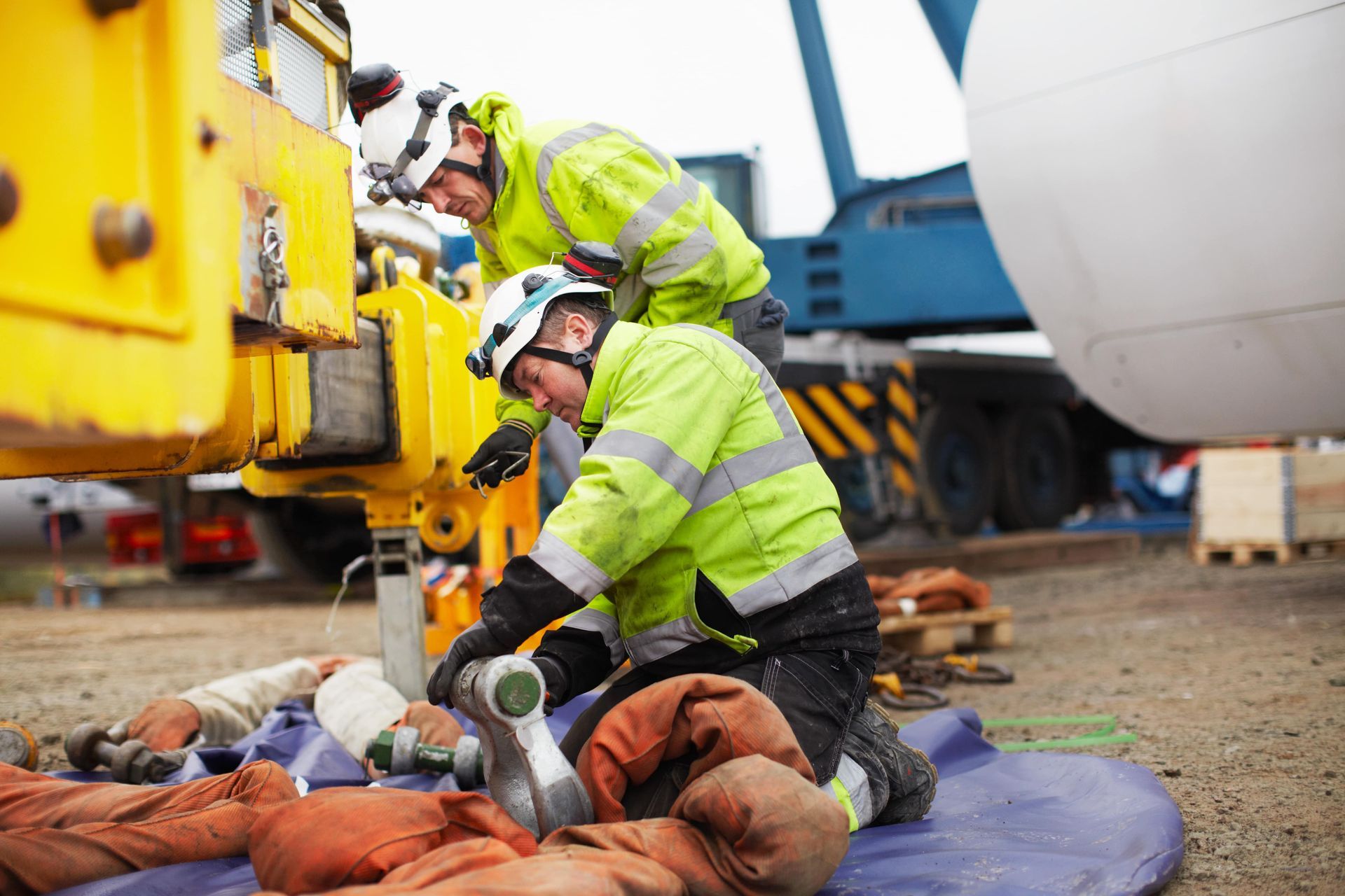 Two construction workers are working on a crane in a parking lot.