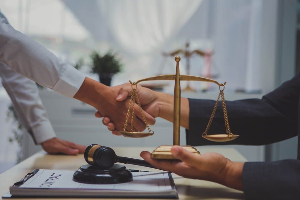 Two people shake hands over a legal contract and a wooden gavel, with a small brass scale of justice held between them.