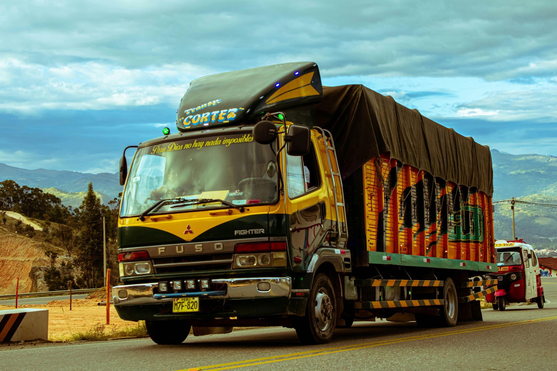 A yellow and dark green Mitsubishi Fuso truck with a black cargo cover travels along a road against a mountainous backdrop.