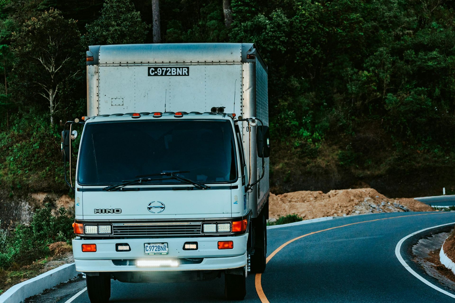 A white box truck driving on a curved, paved road surrounded by lush green trees.