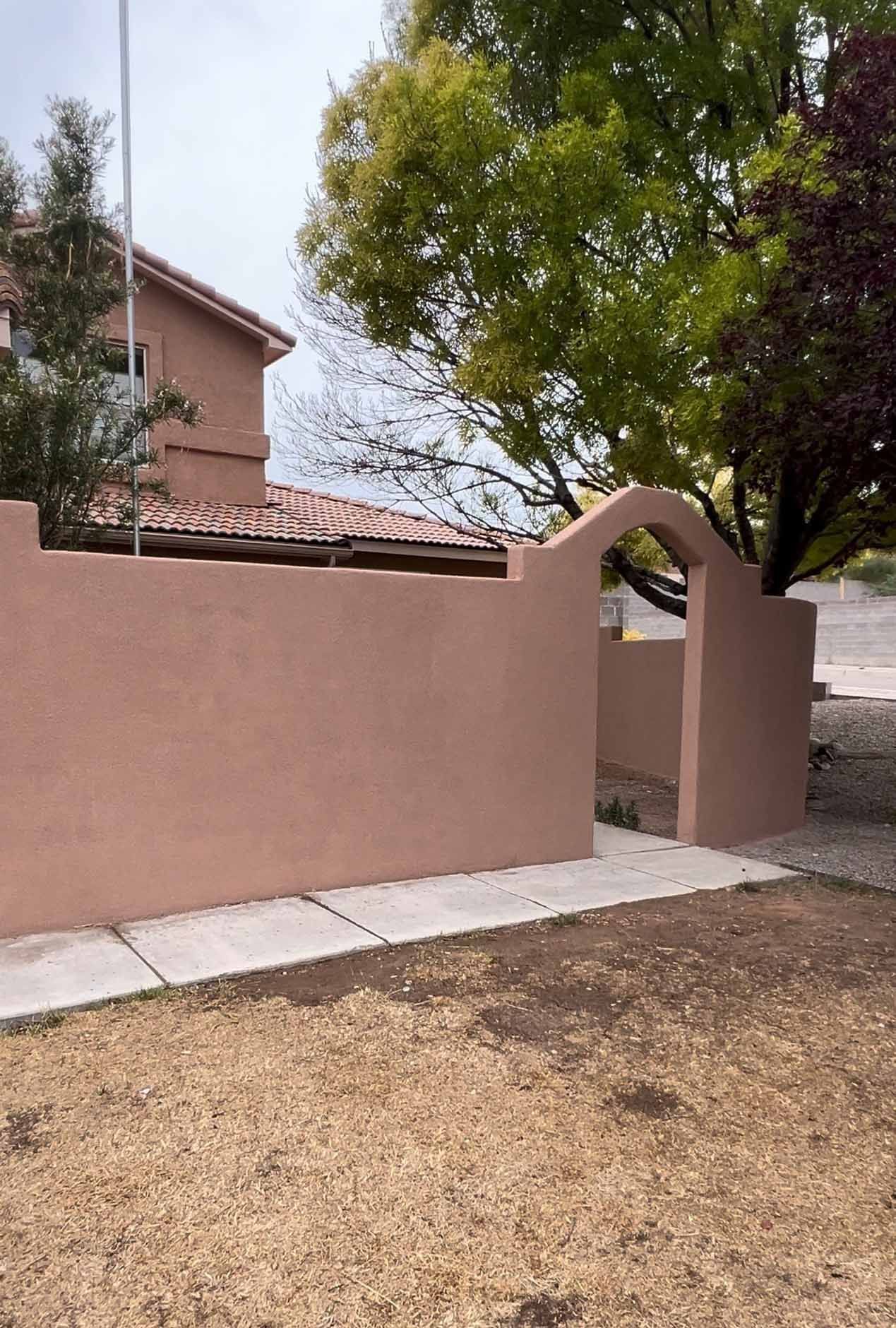 Pink stucco wall with a small arched gate beside a tree and a house in a dry yard