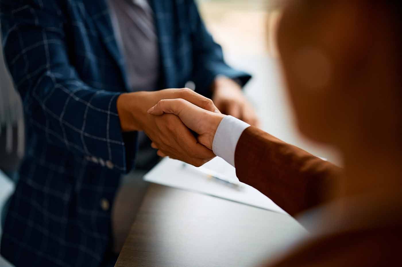 Two people shaking hands over a table, one in a blue plaid jacket and one in a brown sleeve