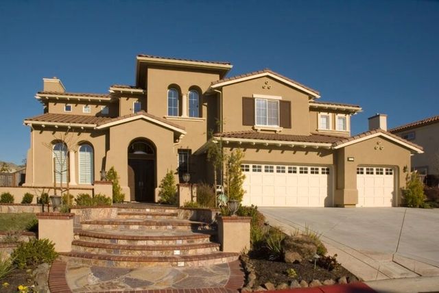 Large tan stucco house with arched entry, tile roof, and a wide driveway under a clear blue sky