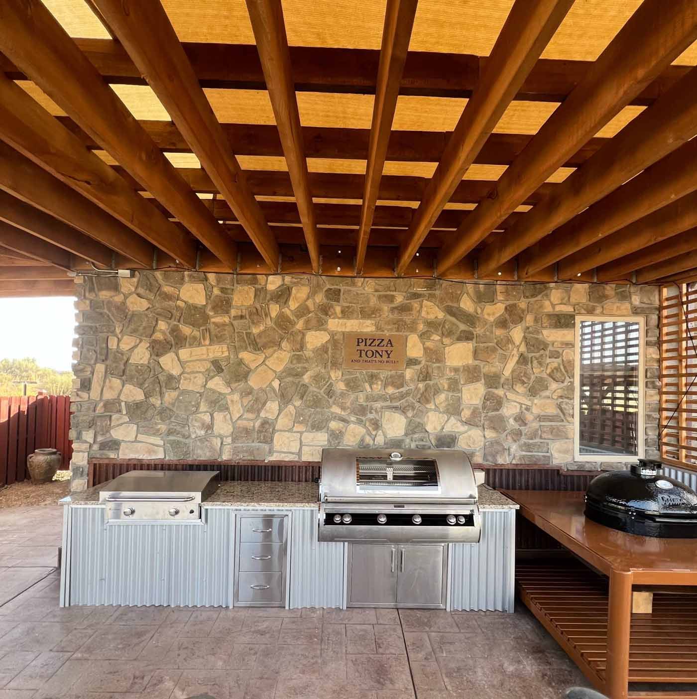 Covered outdoor kitchen with stone wall, wood-beam ceiling, built-in grill, and counter space