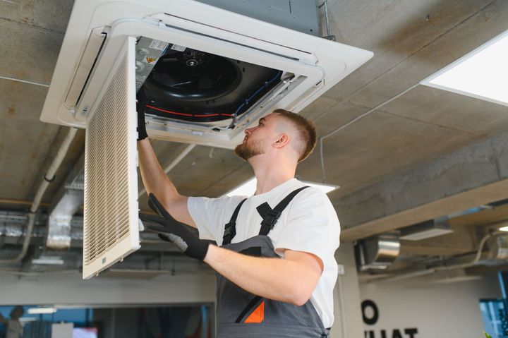 Technician inspecting a ceiling HVAC unit with the access panel open in an indoor industrial setting