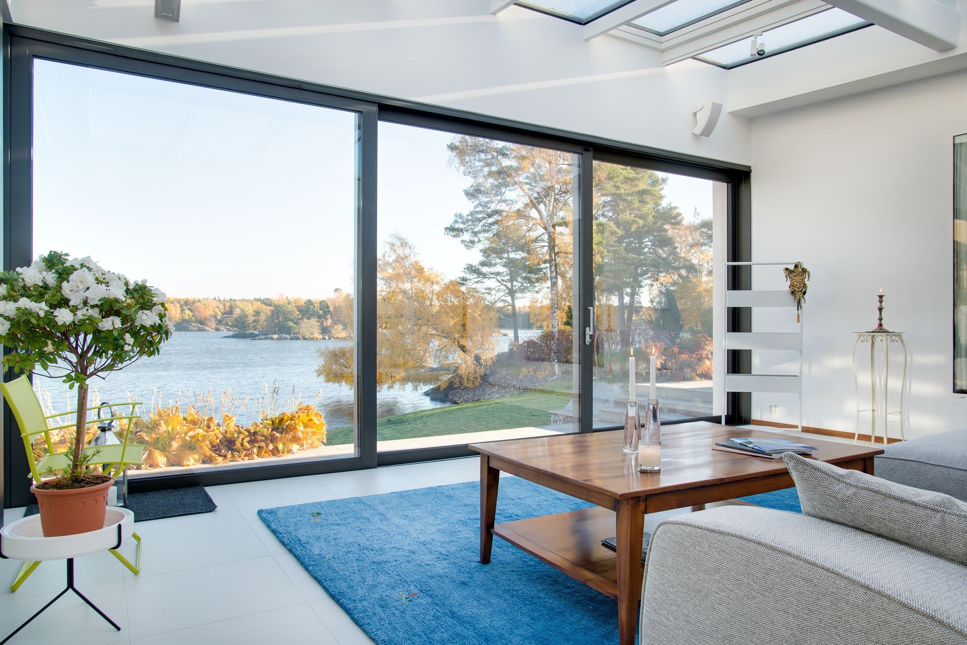 A sunlit living room with large glass sliding doors overlooking a calm lake and autumn trees, featuring a blue rug.