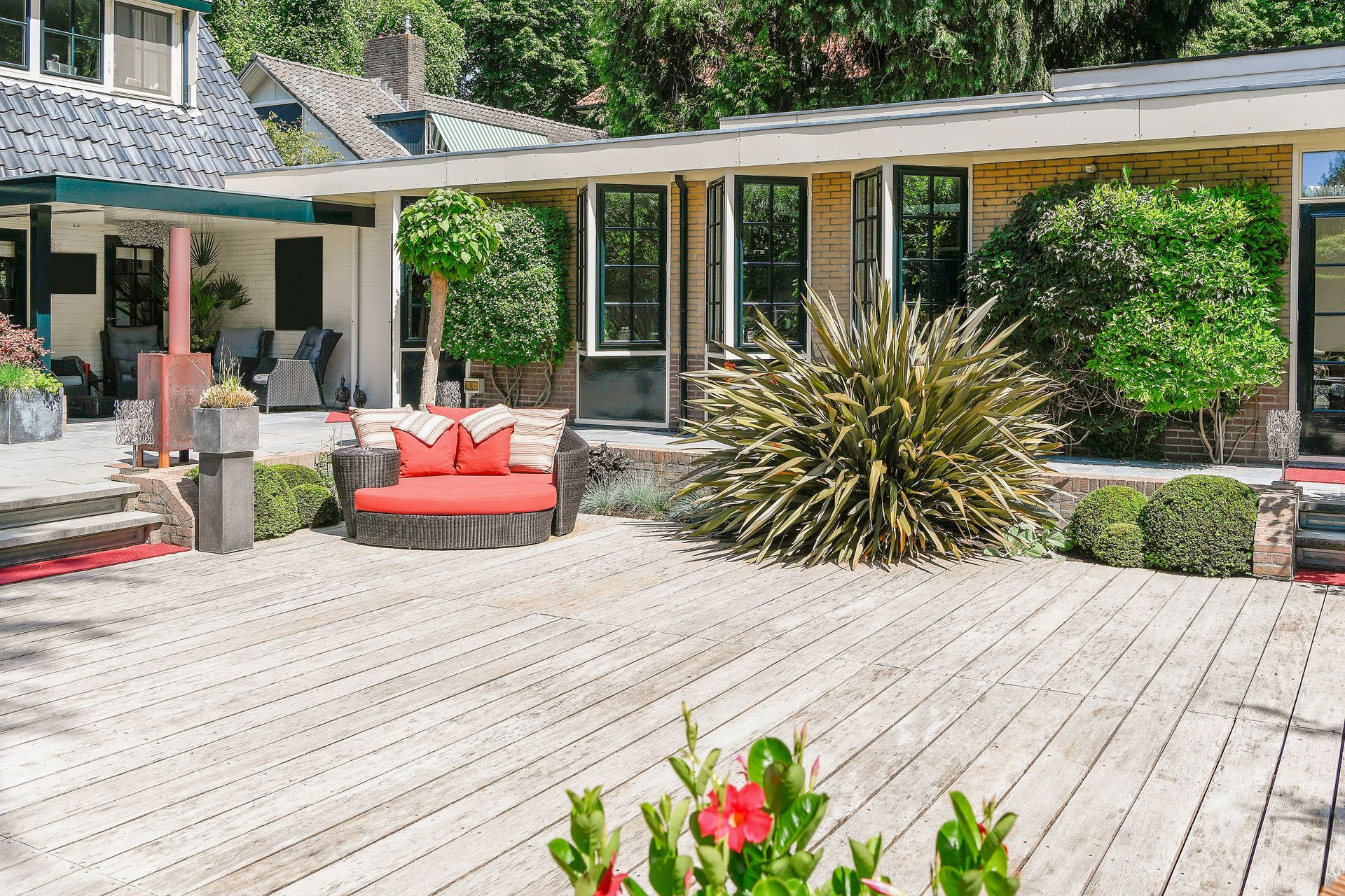 A wooden deck with a round red outdoor sofa and green plants in front of a house with large windows and a garden.