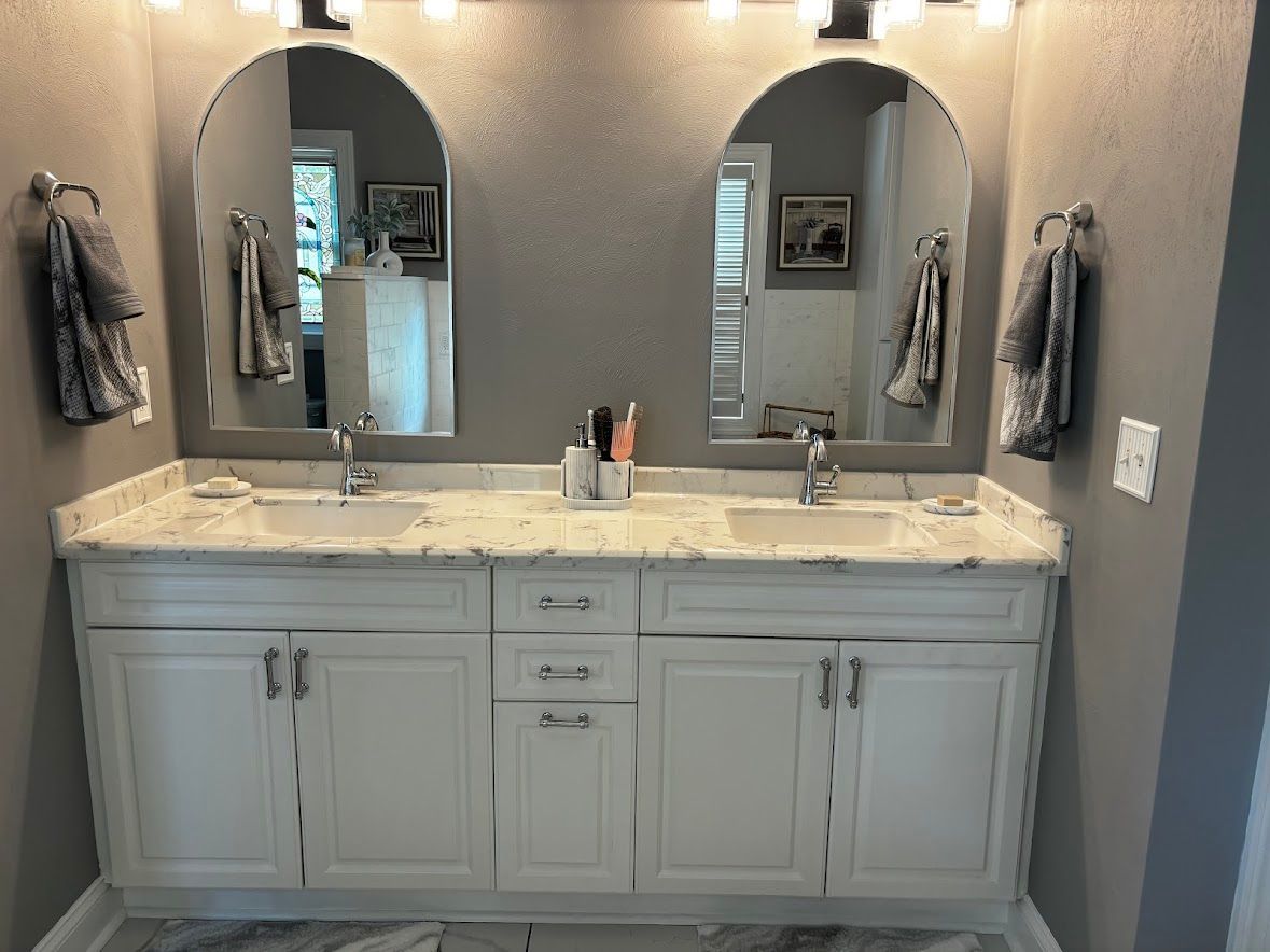 Double vanity with marble countertop, white cabinetry, two arched mirrors, and grey walls in a bathroom.