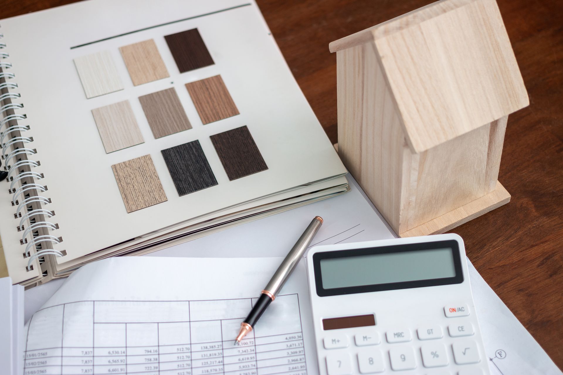 A wooden house is sitting on a table next to a calculator and a pen.