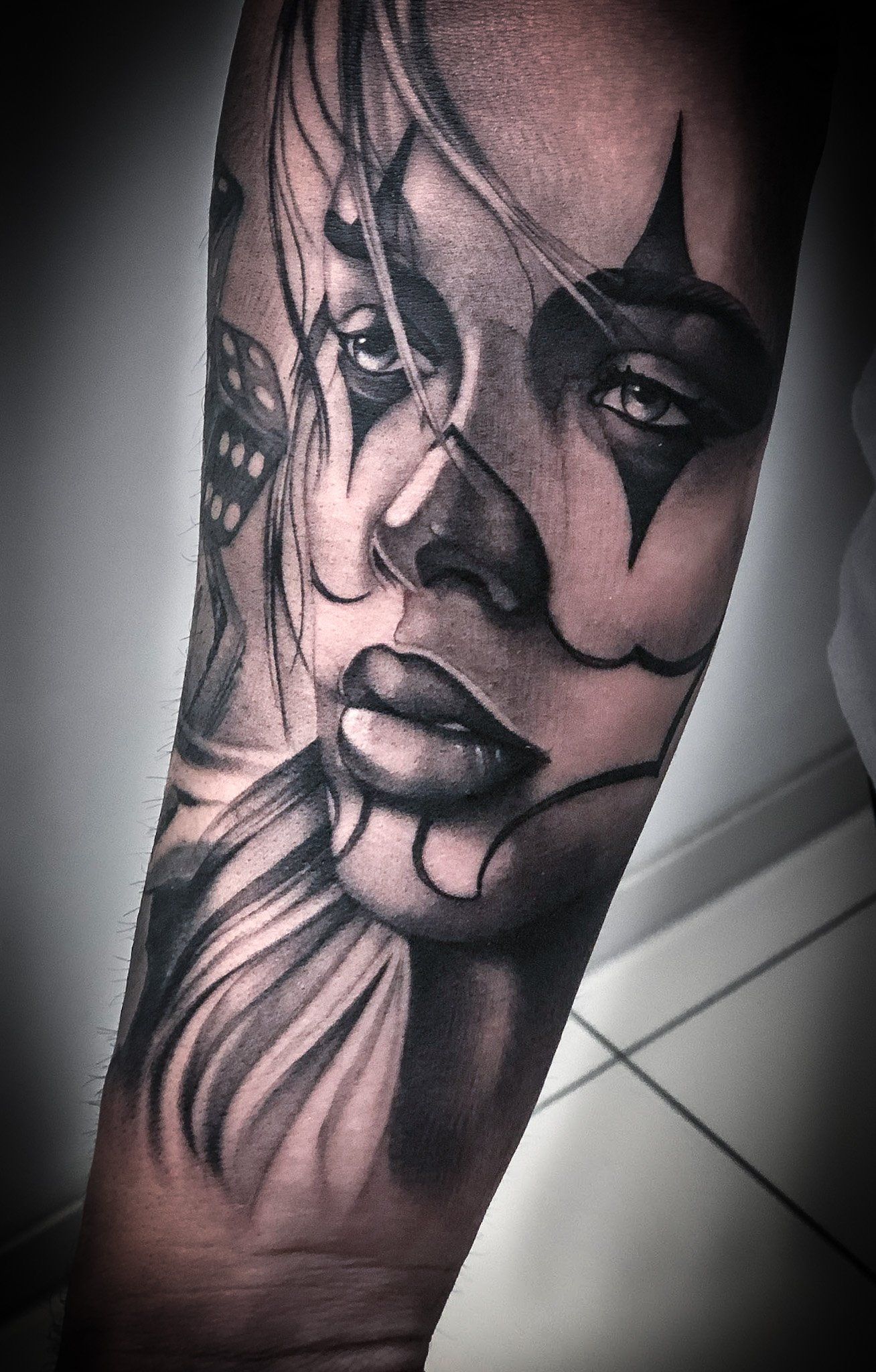 Ein schwarz-weißes Tattoo einer Frau mit einem Clownsgesicht auf dem Arm.