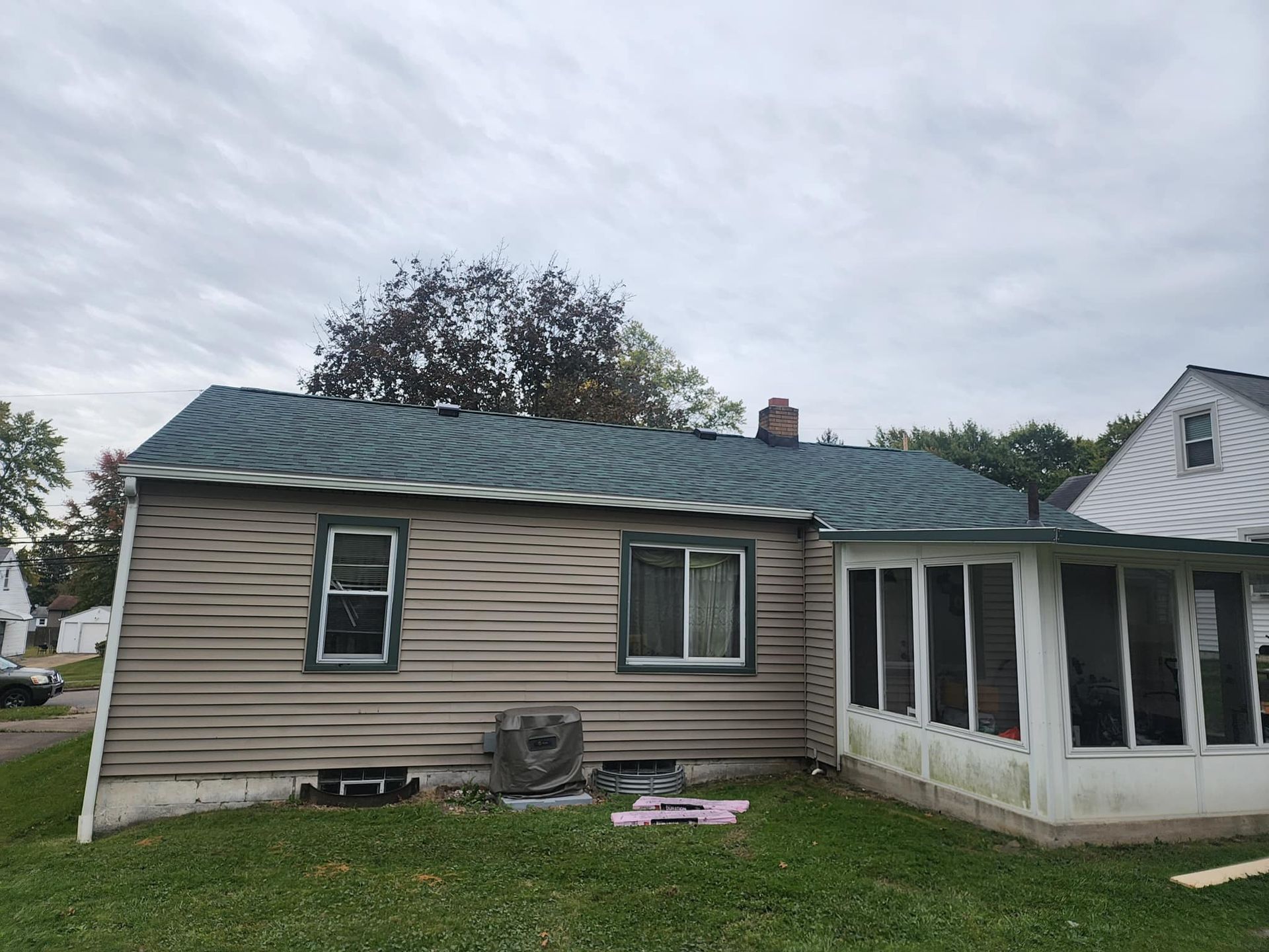Beige house exterior with green roof and enclosed porch, viewed from the backyard under a cloudy sky
