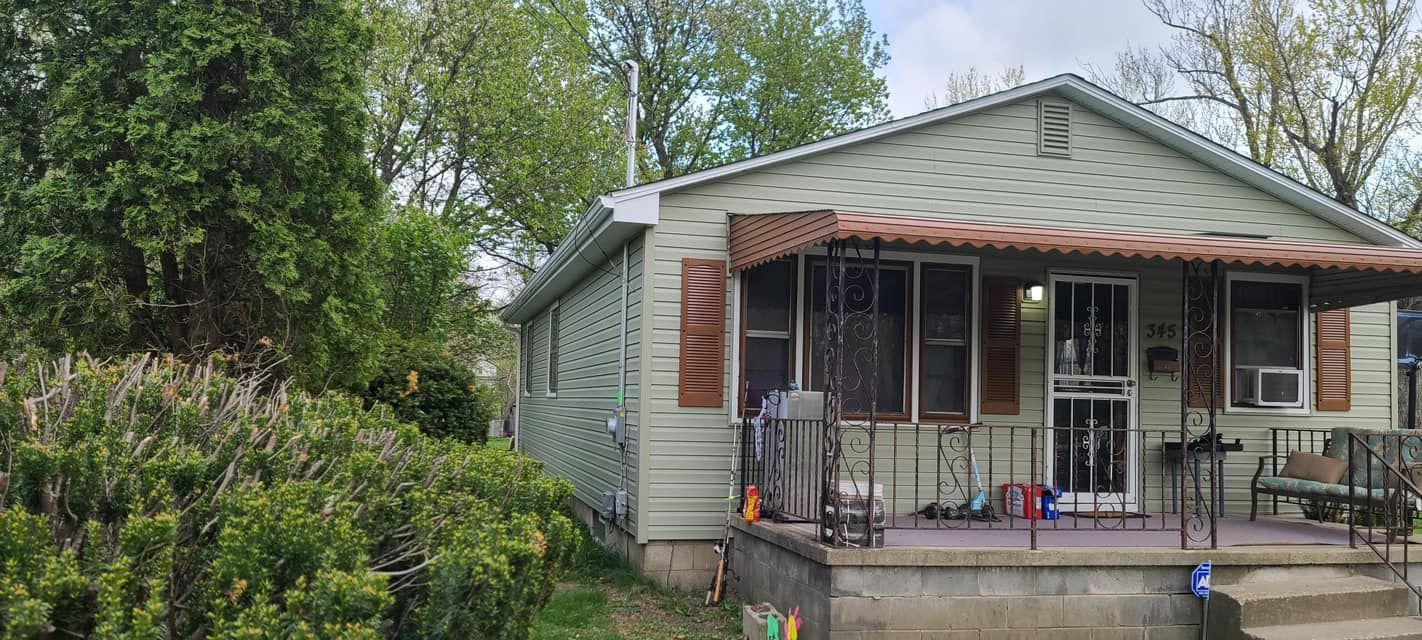 Small house with a porch, red shutters, and greenery in the yard after rain
