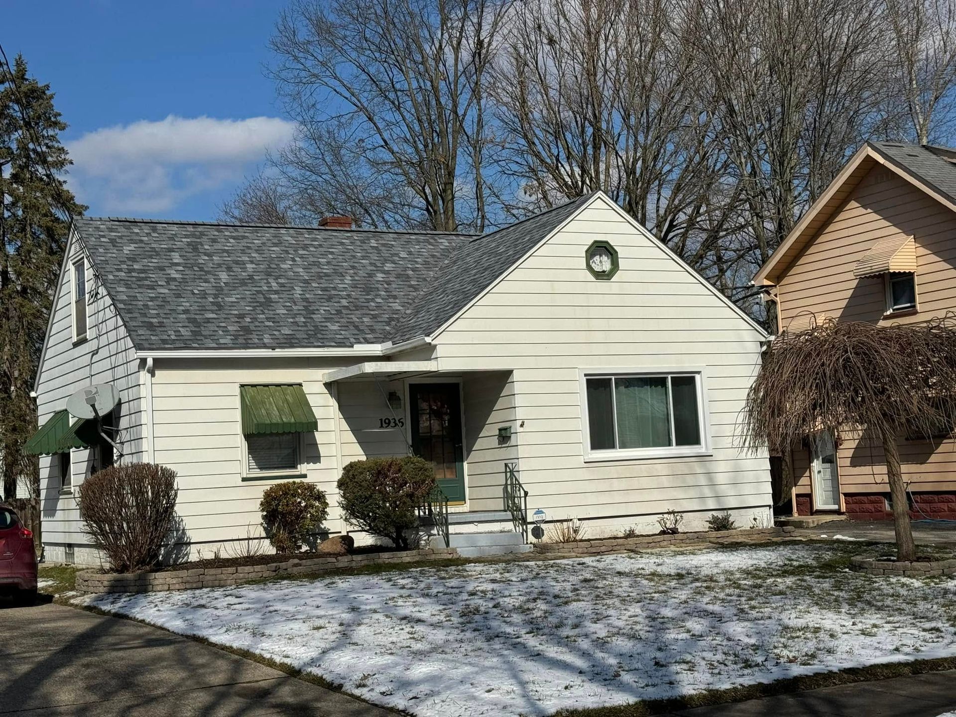 Single-story white house with green awning, snow-dusted lawn, and bare trees under a blue sky.