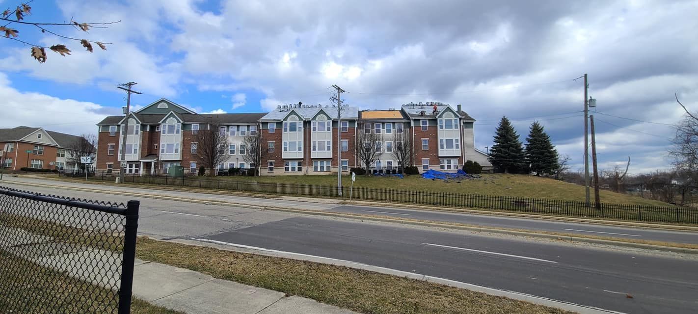 Multi-story apartment complex behind a road and fence under a cloudy sky