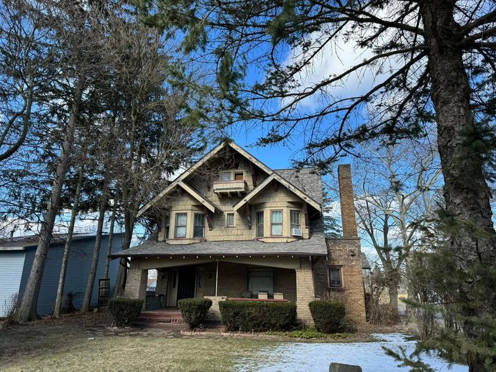 Two-story house with a front porch, stone chimney, and bare trees under a blue sky