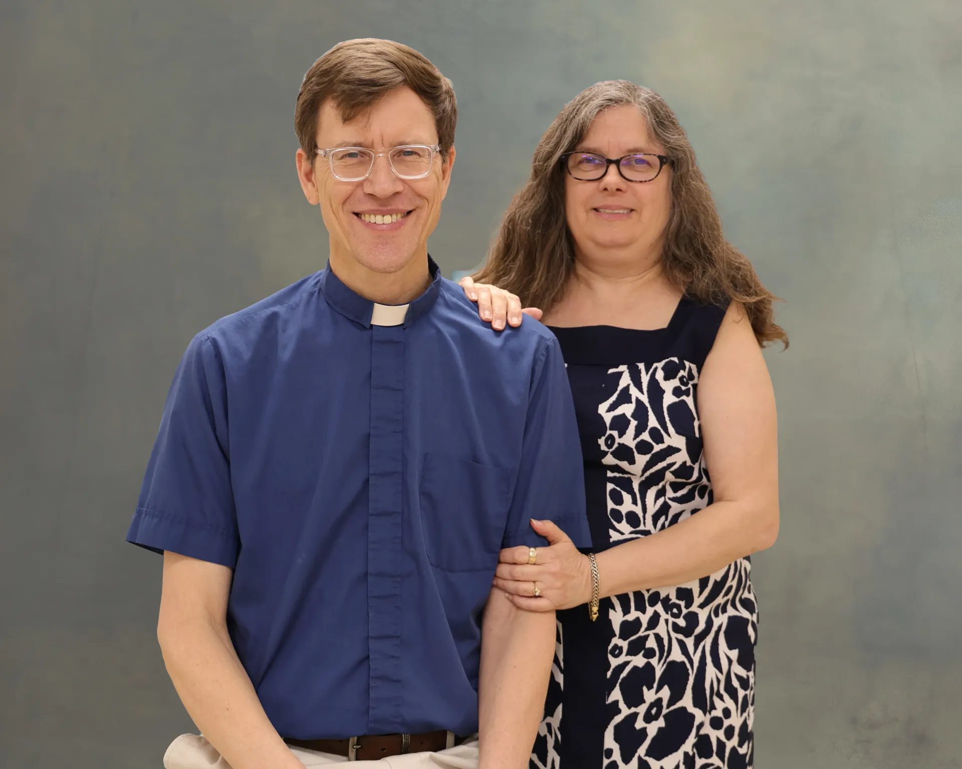 Two people posing against a gray backdrop, smiling, one in a blue clergy shirt and one in a black-and-white dress.