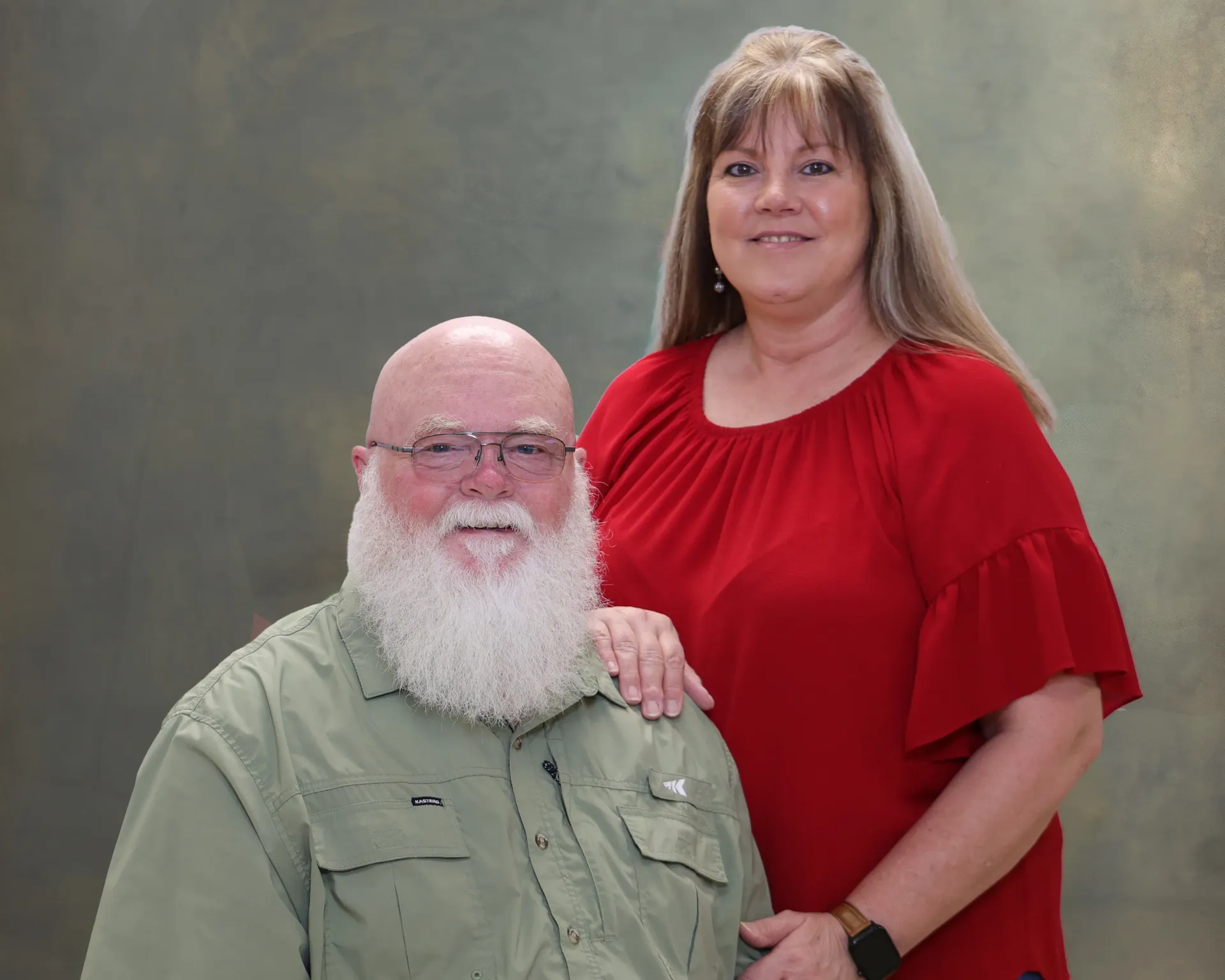 Two people posed in a studio portrait, smiling; one seated in a green shirt, one standing in a red dress.