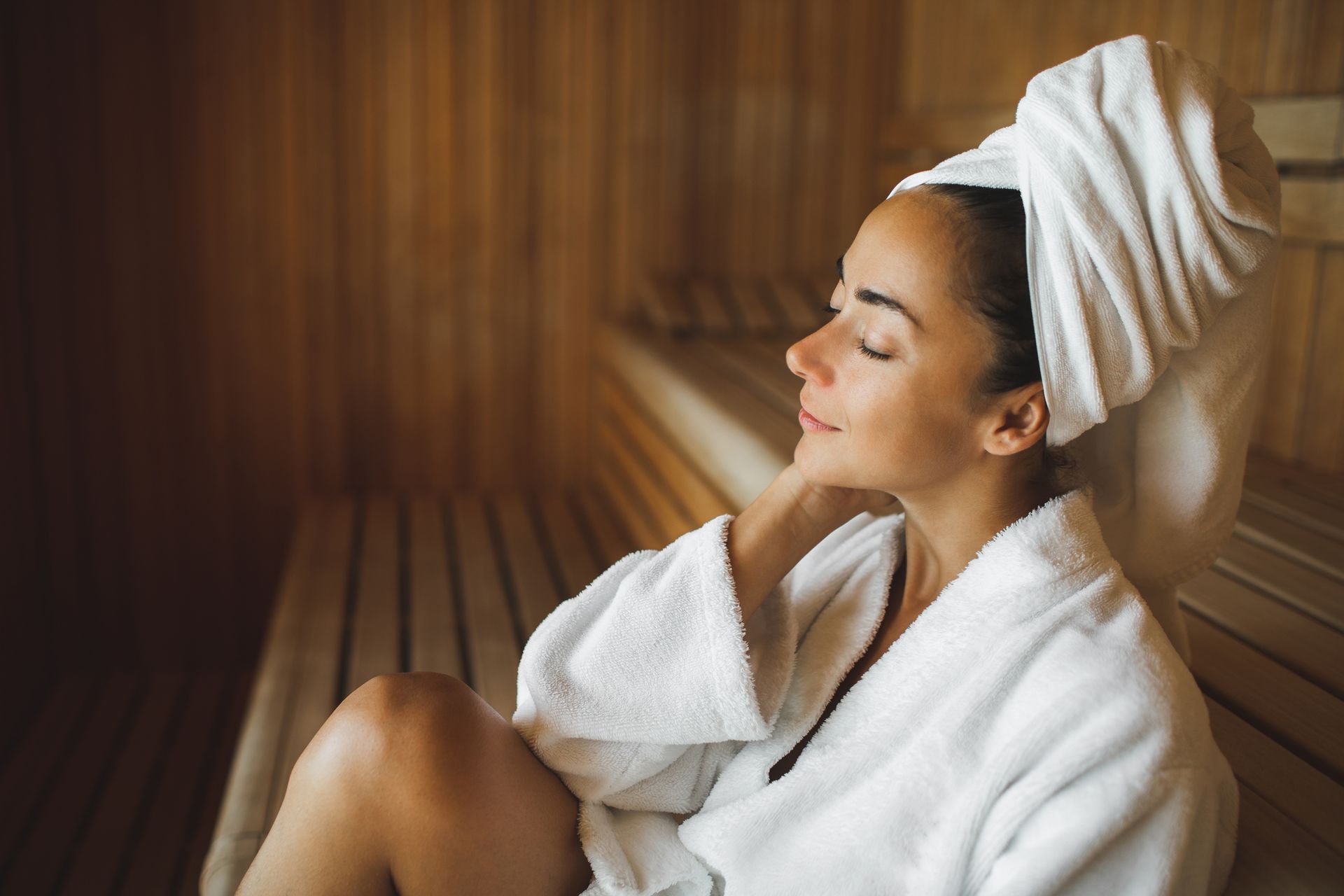 Donna che si rilassa in una sauna, avvolta in un asciugamano e un accappatoio, con gli occhi chiusi. Interni in legno, luci soffuse.
