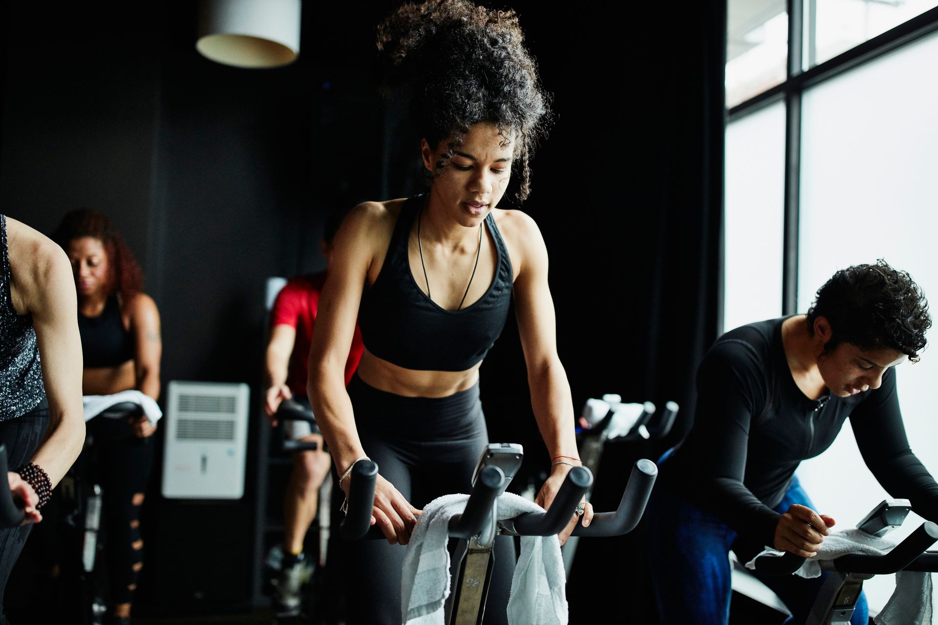 Persone che pedalano durante una lezione di spinning. Una donna in abbigliamento sportivo nero pedala, concentrata. Altri pedalano sullo sfondo. Interno buio.
