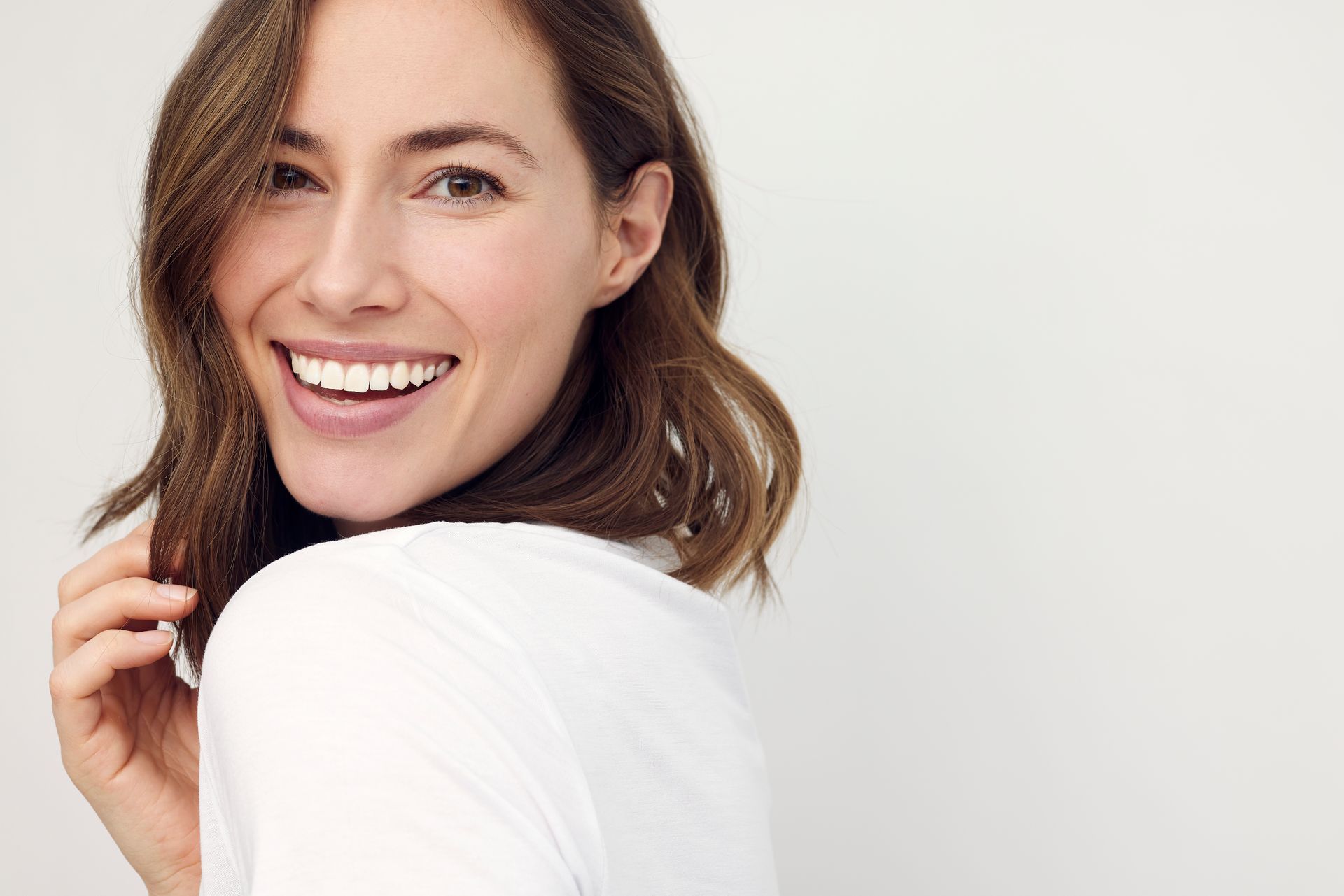 Woman with short, wavy brown hair smiles at the camera, wearing a white shirt against a white backdrop.