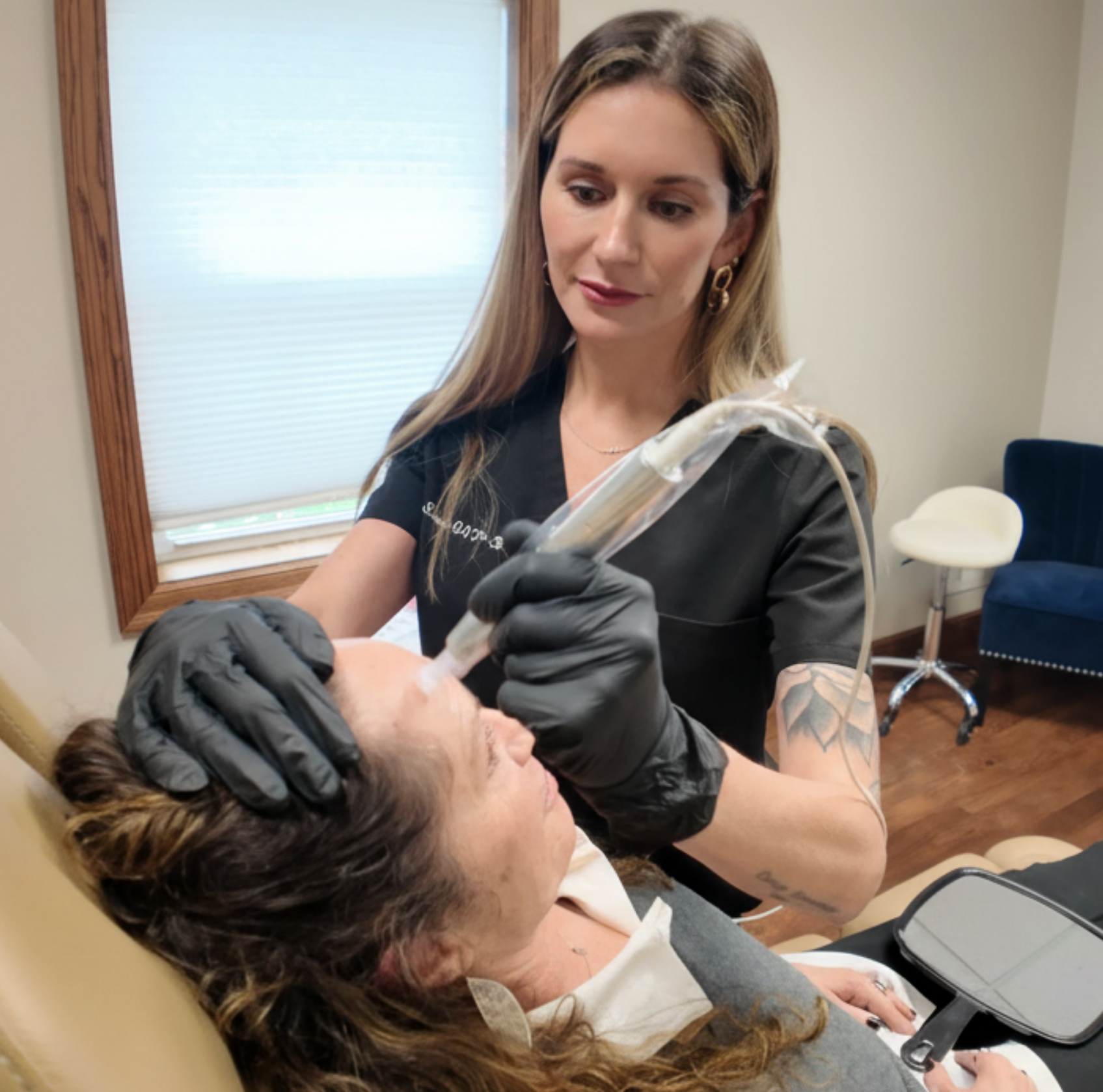 Woman performs a cosmetic procedure on a client's face with a pen-like device. Both wear black gloves, in a salon setting.