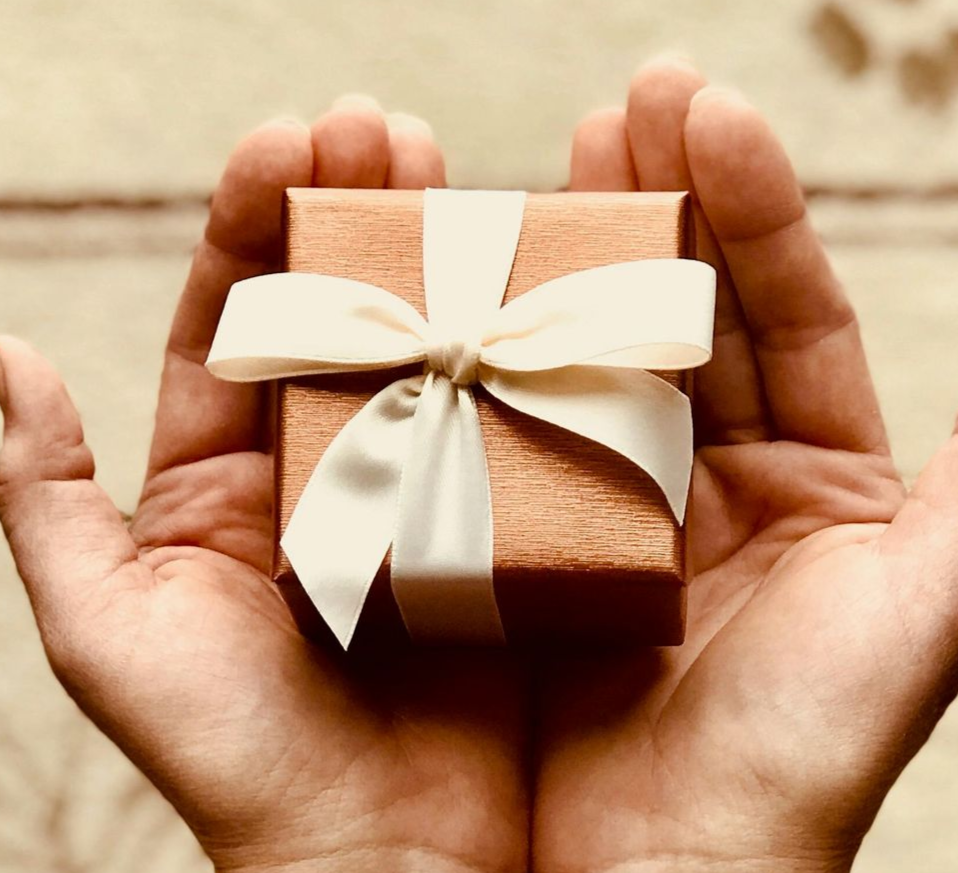 Hands cupped, holding a small, square, gold gift box with a white ribbon.