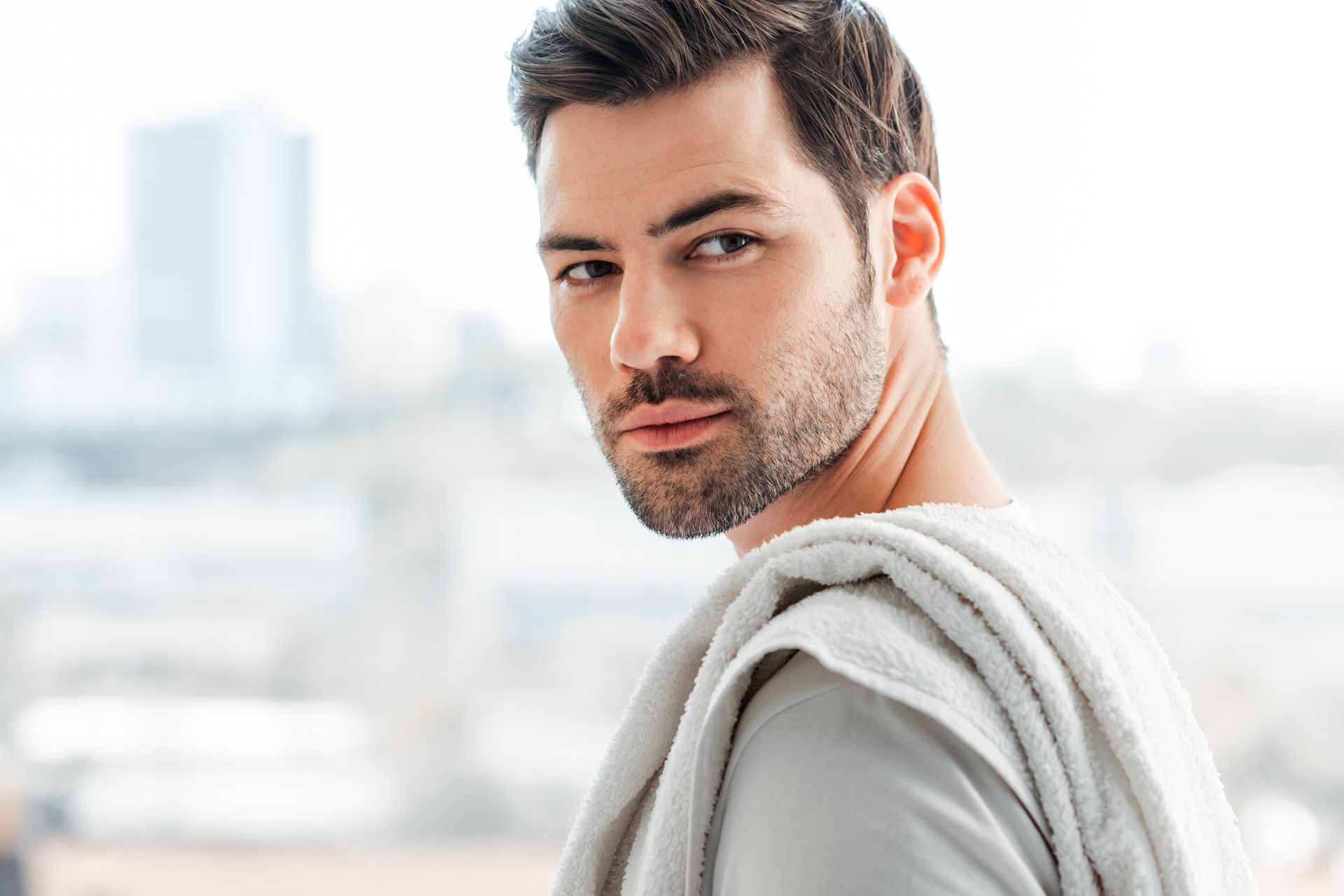 Man looking over shoulder with towel; light-colored shirt, blurred city backdrop.