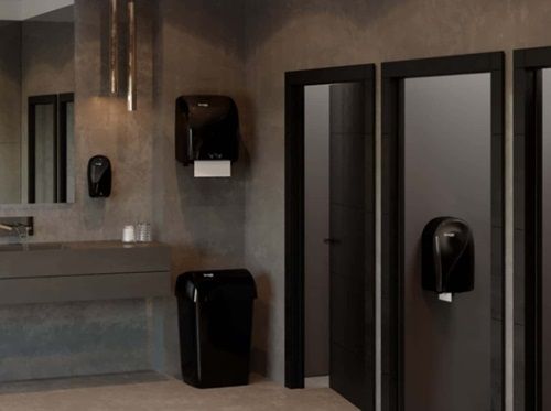 Public restroom with black paper towel dispensers, doors, and trash can against a grey wall.