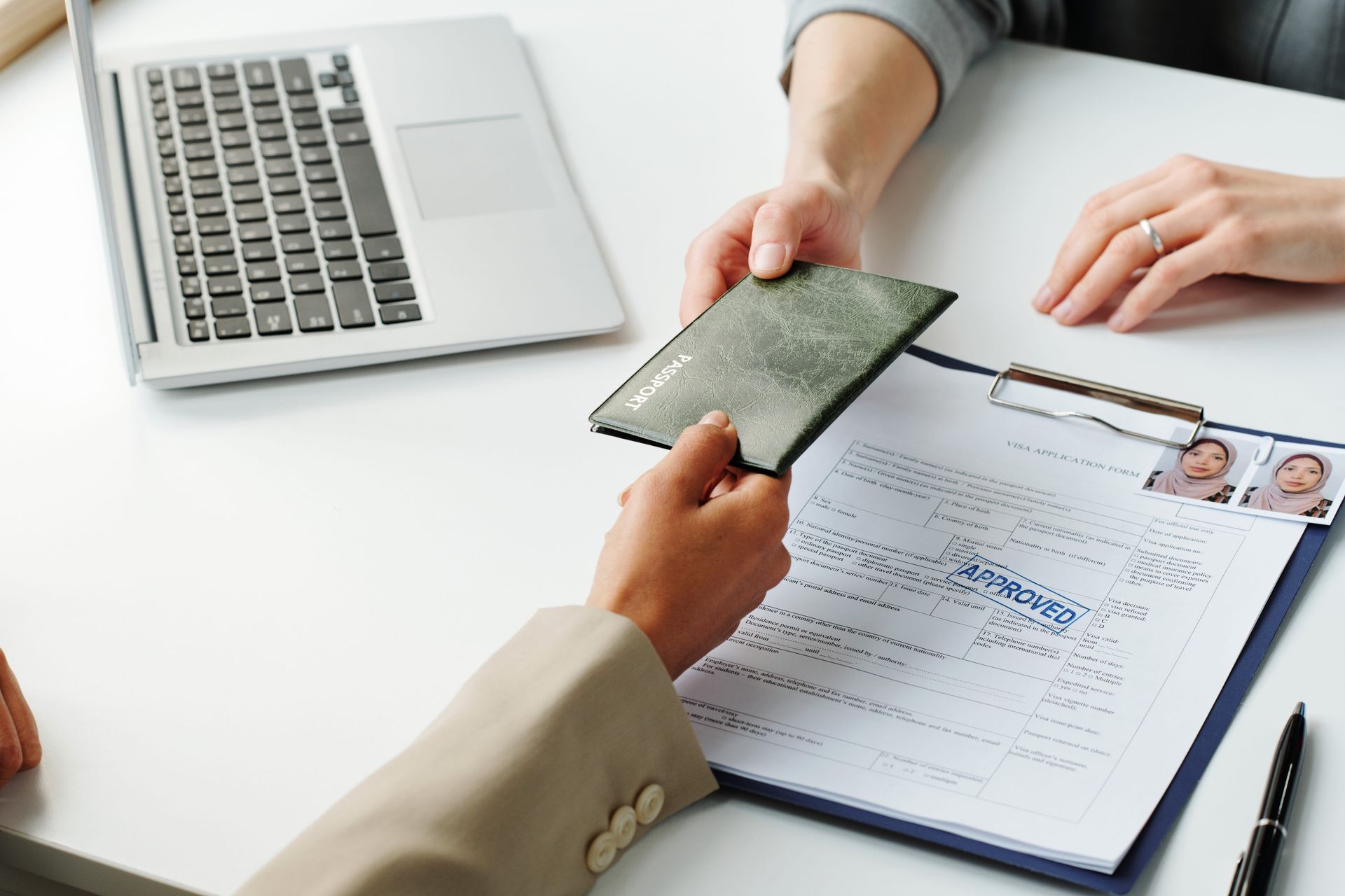 Hands exchanging a wallet over a desk with forms, a laptop, and ID photos visible