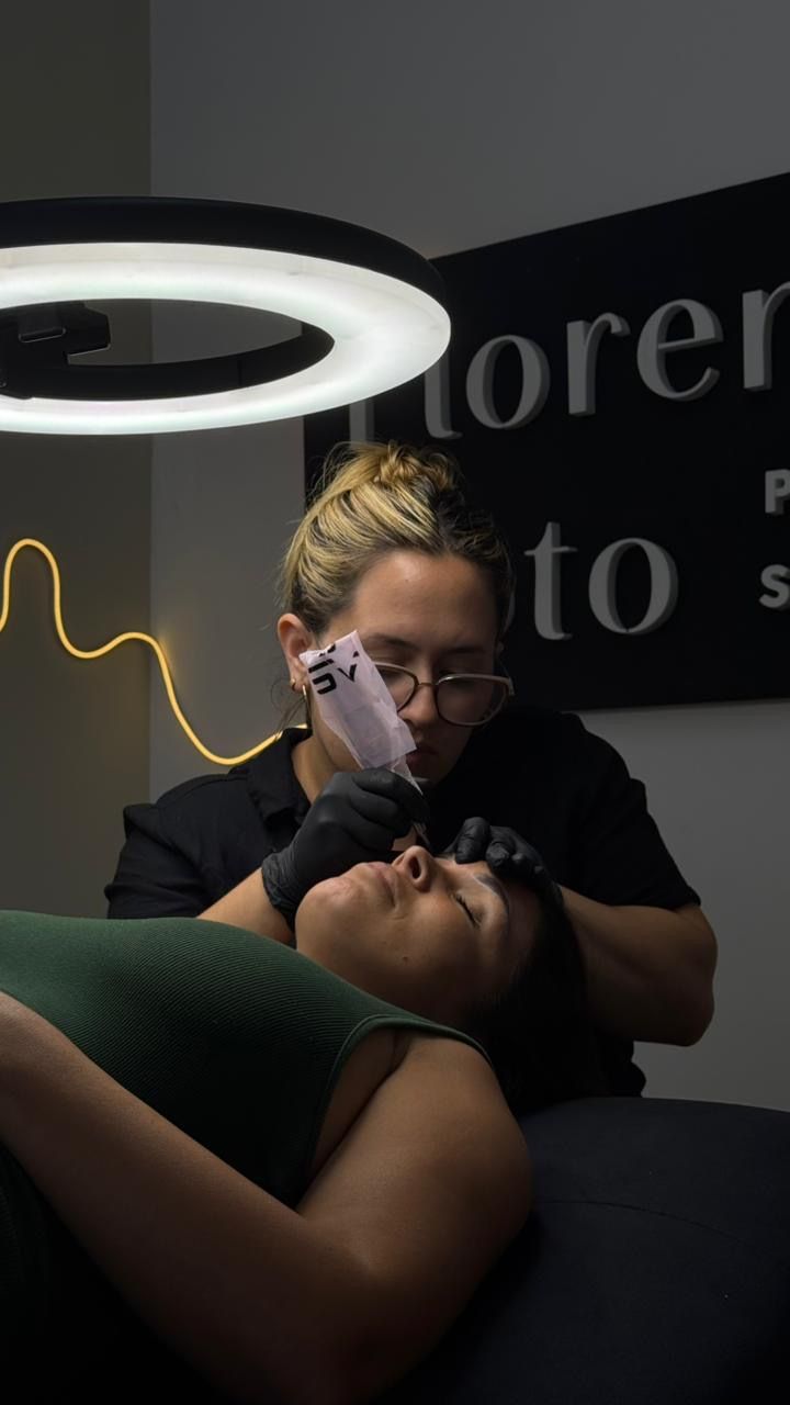 Una profesional realiza un procedimiento cosmético de cejas a una clienta que está tumbada bajo una luz anular en un estudio.