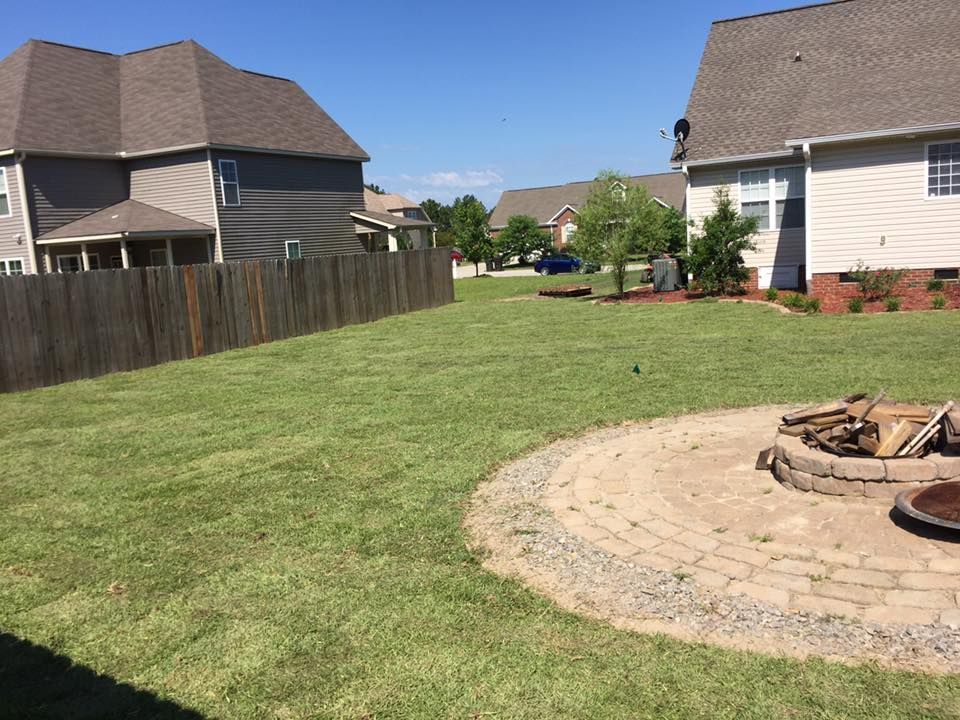 Backyard with green sod lawn, wooden fence, houses, and a circular stone fire pit patio in Pittsboro, NC