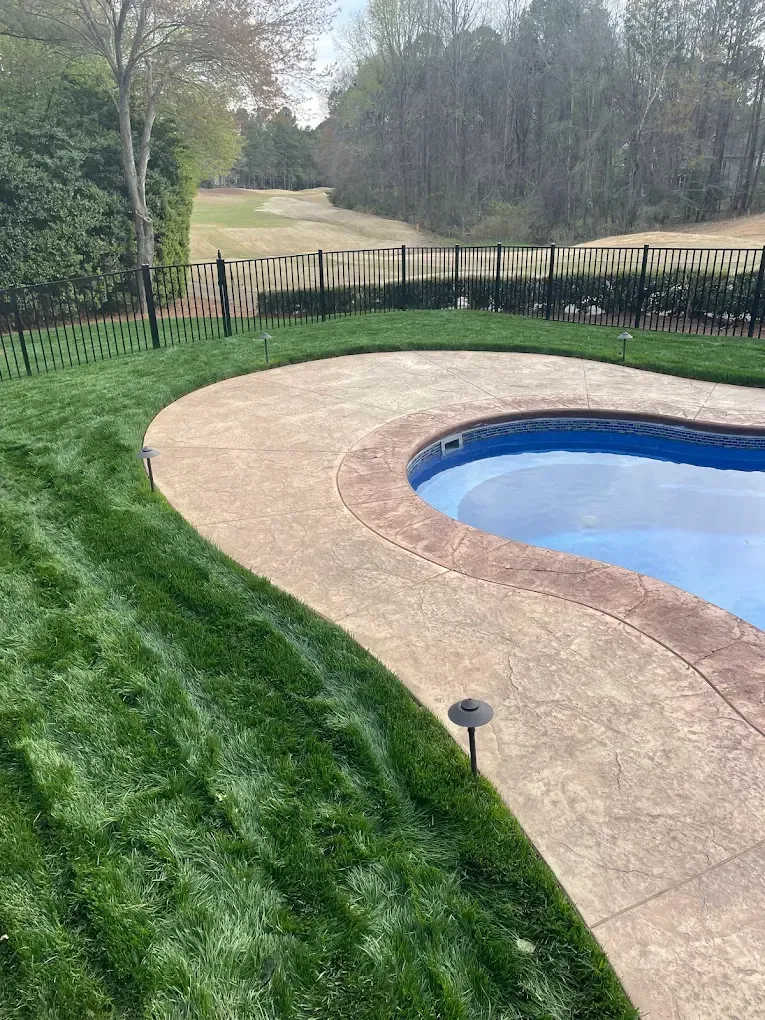Large grassy backyard with a brick house, covered porch, and trees along the fence line.
