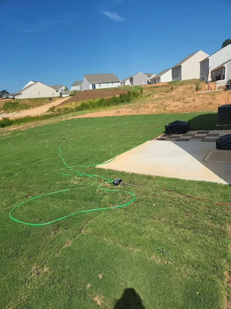 Backyard lawn beside a concrete patio, with a green hose and suburban houses on a hillside.