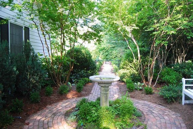 Brick garden path leading to a white stone birdbath amid lush green shrubs and trees