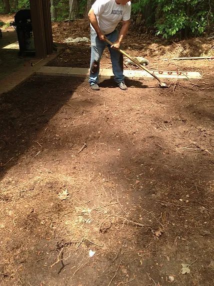 Person raking a dirt path outdoors near a house and trees.