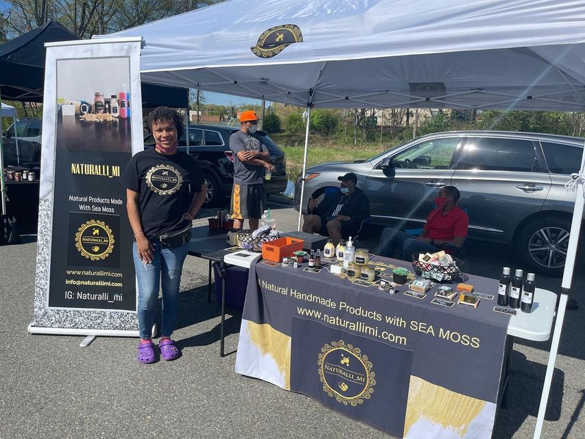 Woman at a booth selling products labeled "Wellness with Sea Moss". Others nearby. Sunny outdoor setting.