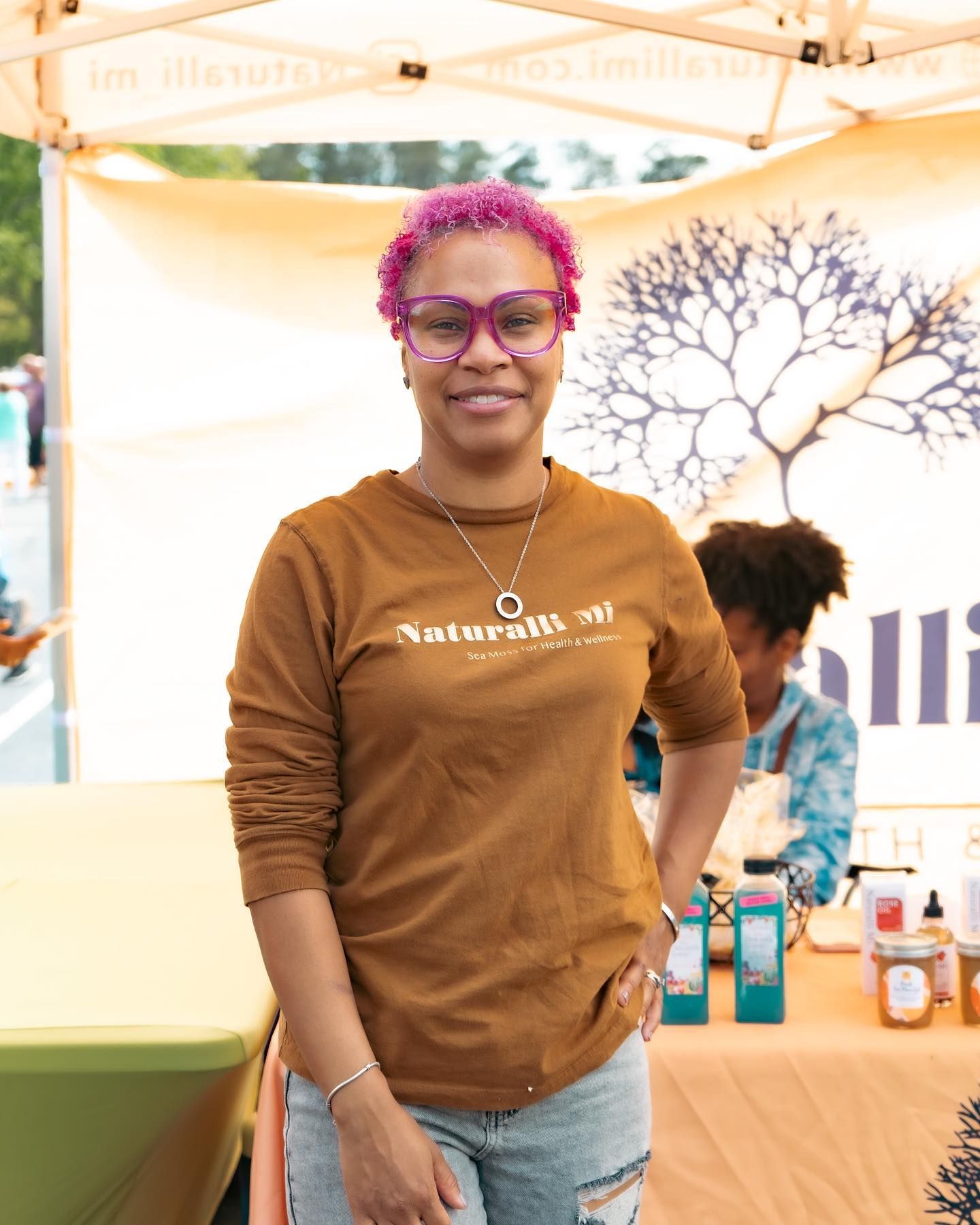 Woman with pink hair and glasses stands at a vendor booth, smiling. She's wearing a brown shirt and jeans.