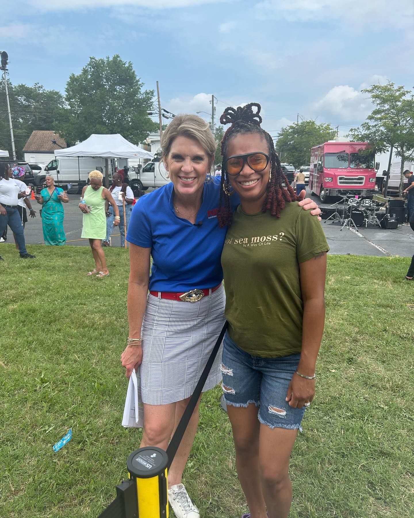 Two women smiling and posing for a photo outdoors in a park. One in a blue shirt and skirt, the other in a green shirt.