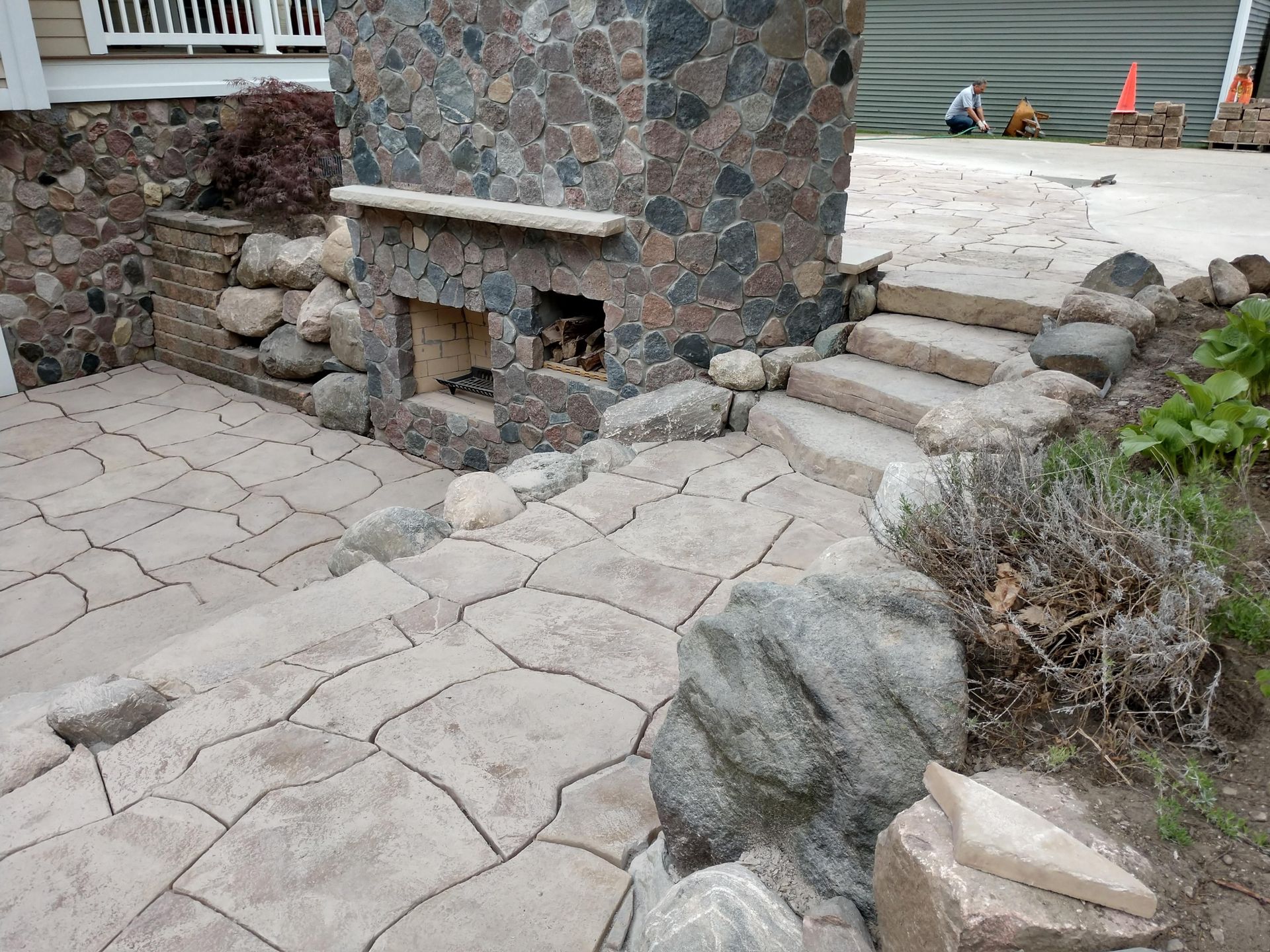 A stone patio featuring a stone fireplace and steps leading to a higher concrete level, surrounded by landscaping rocks.