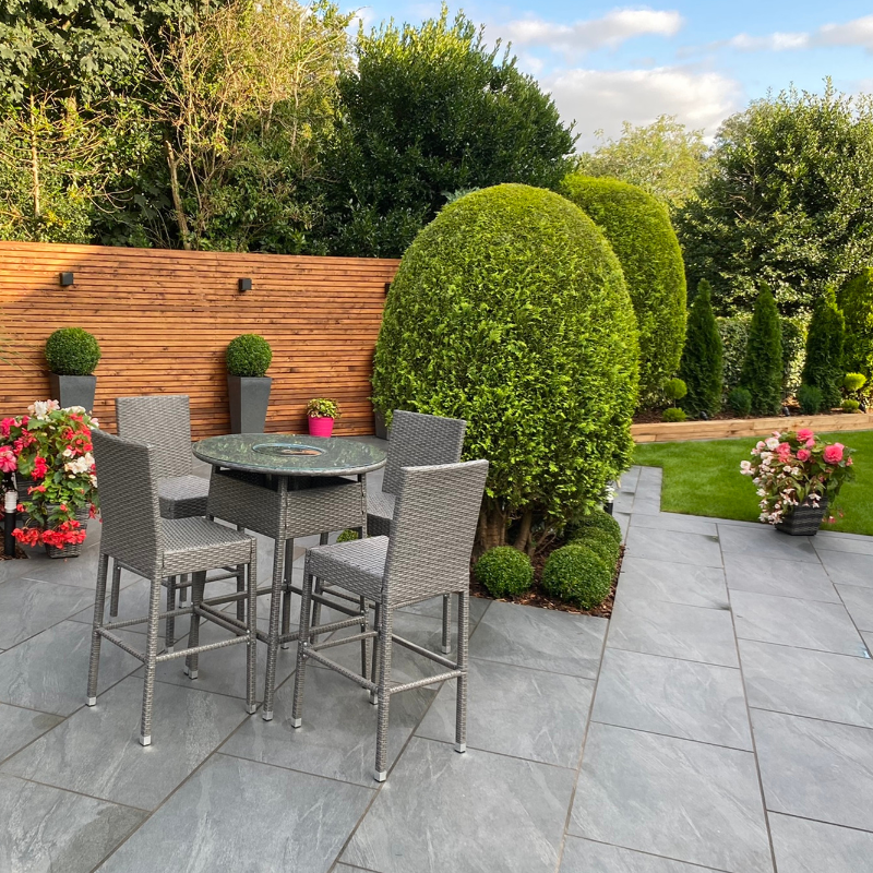 Patio with gray chairs, table, and tall green bushes. Wooden fence and flower pots in a sunny garden.
