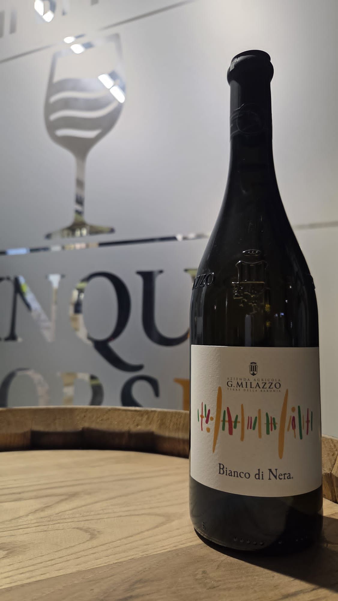 Bottiglia di vino su una superficie di legno, con una sagoma di vetro e un logo sullo sfondo.