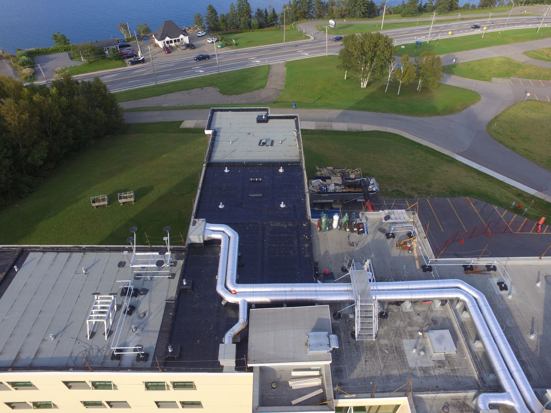 Aerial view of a building rooftop with silver ducts, parking lot, and surrounding roads and grassy areas