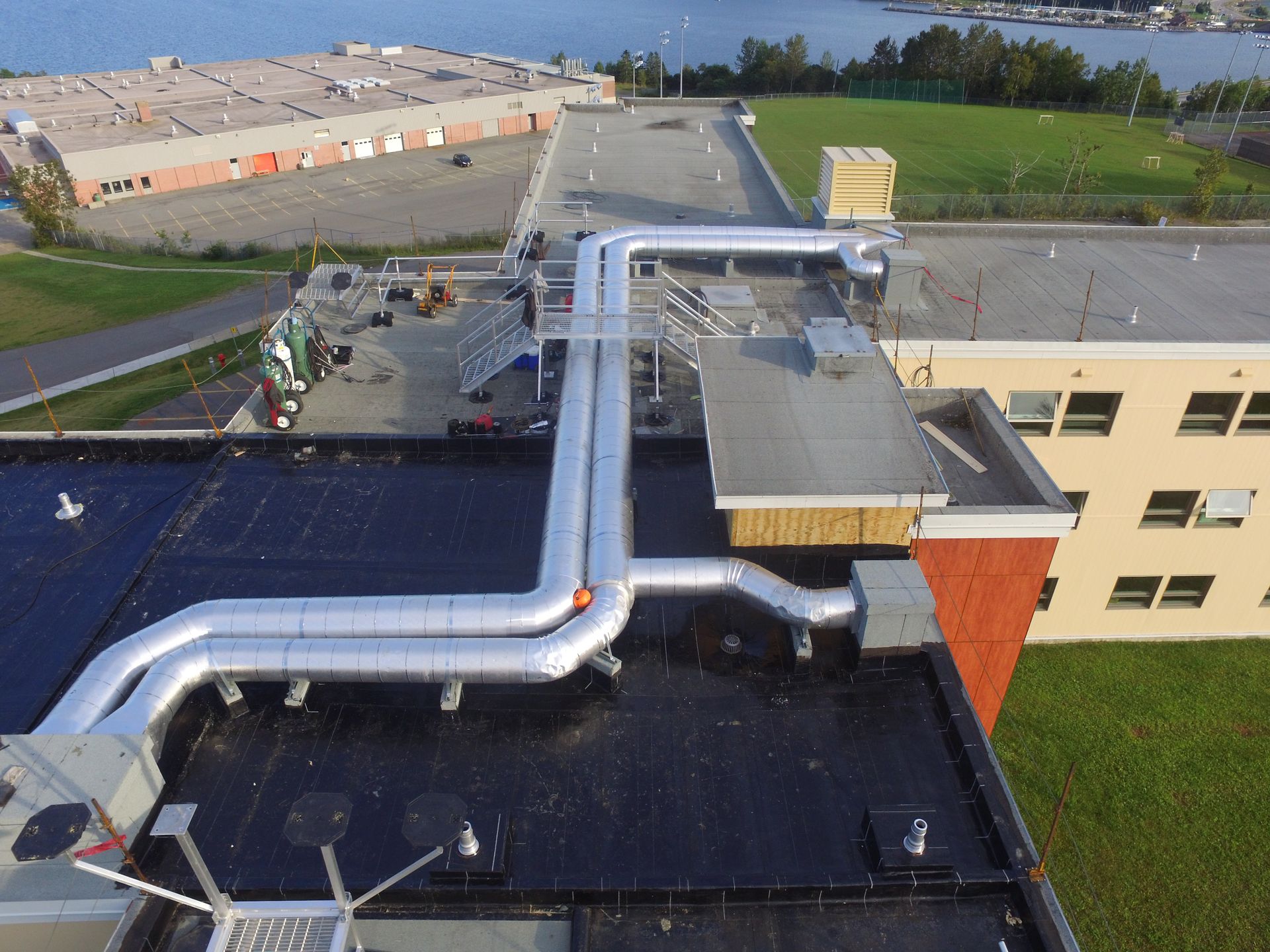 Rooftop with silver HVAC ducts and black roofing beside a beige building and green field