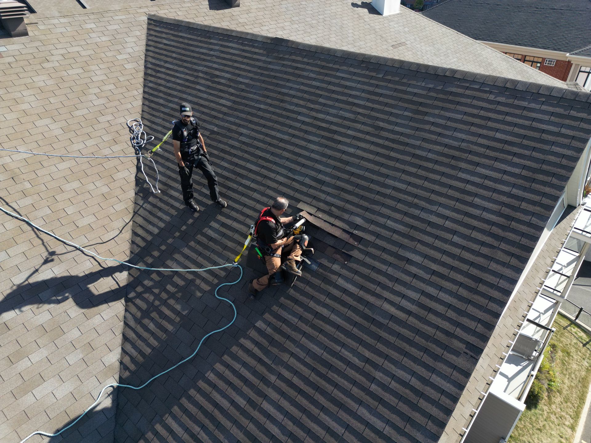 Two roof workers on a dark shingled roof, one standing and one kneeling beside safety ropes.