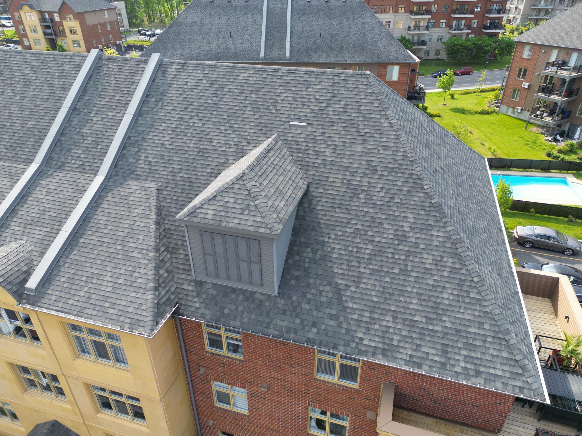 Aerial view of a gray-shingled rooftop on a brick apartment building with surrounding buildings and trees.