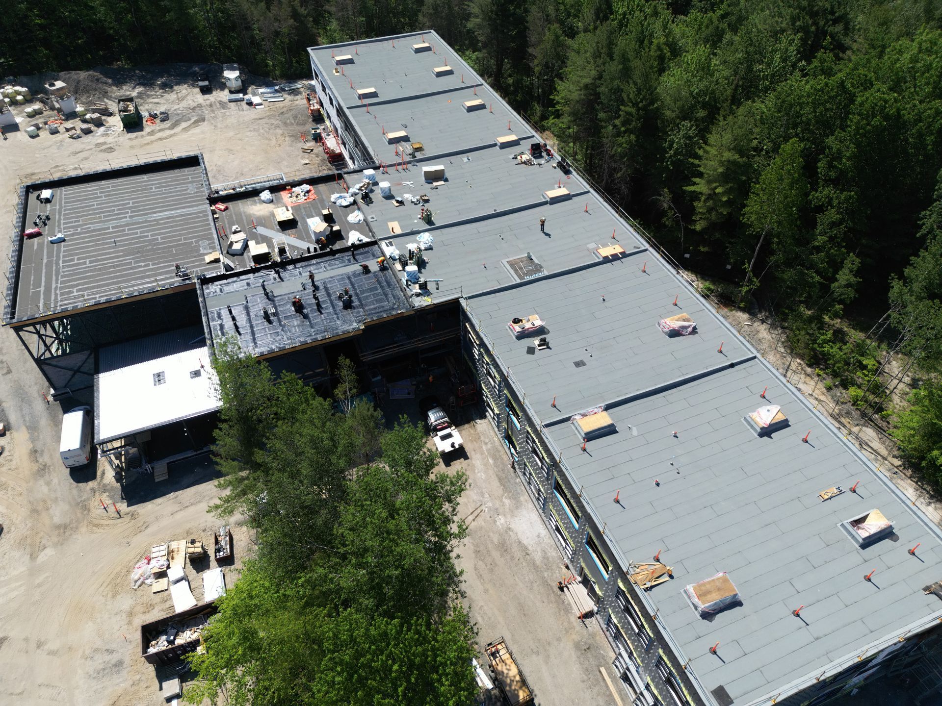 Aerial view of a long gray-roofed industrial building beside trees and a paved yard.