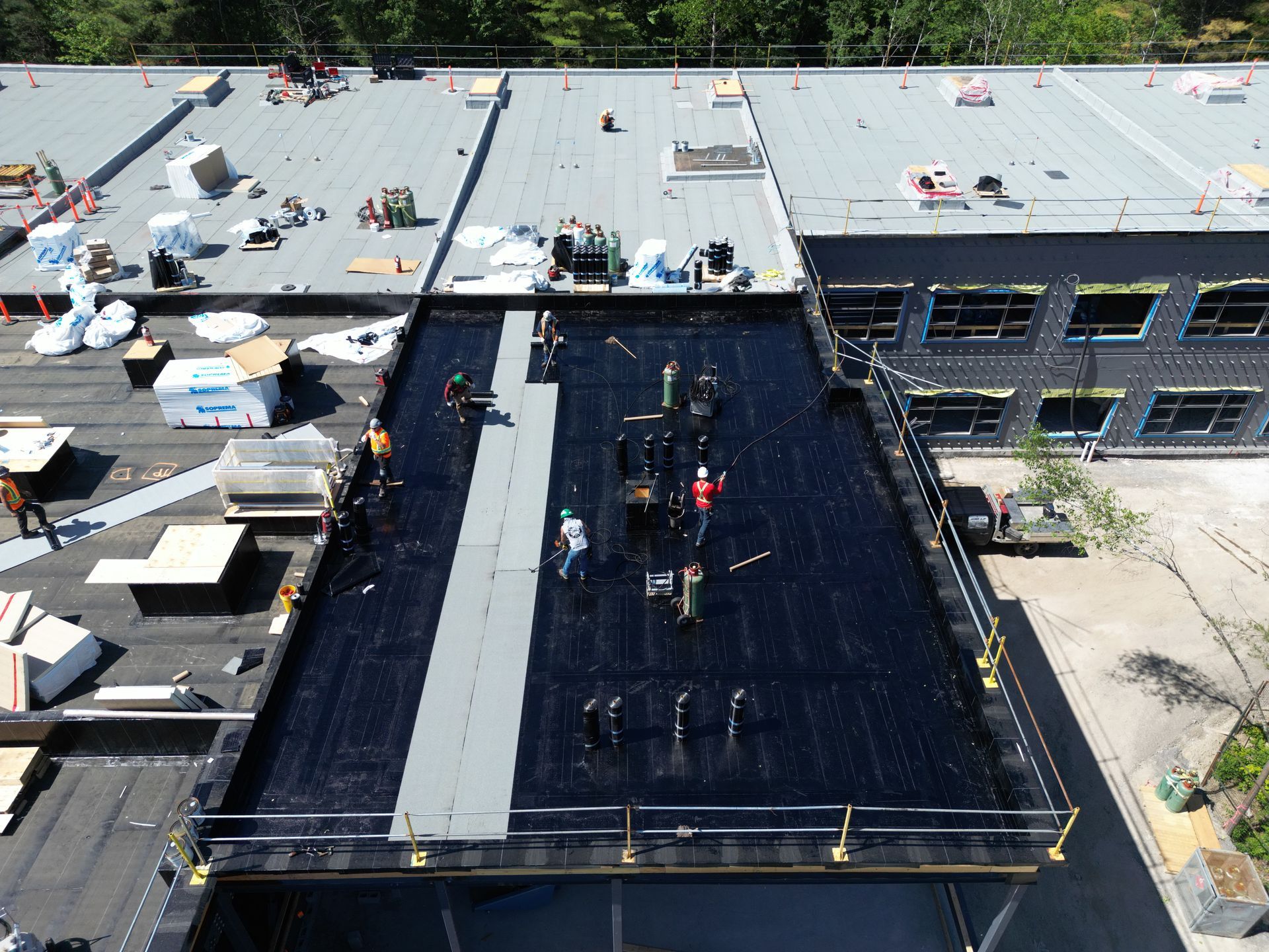 Aerial view of rooftop workers on a black roof between two white buildings, with equipment and materials scattered around.