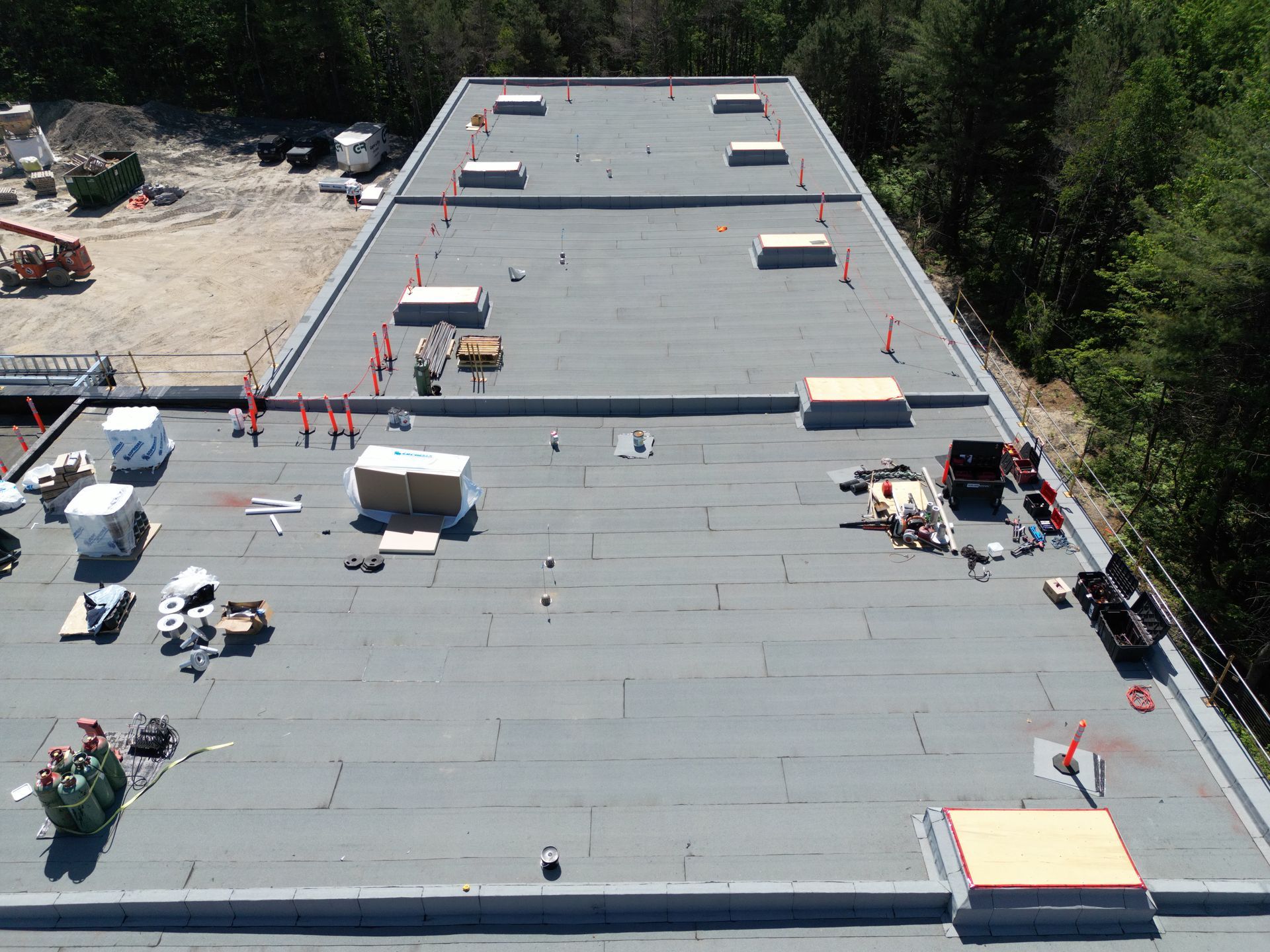 Aerial view of a flat roof with construction materials, workers, and orange safety barriers near a wooded area