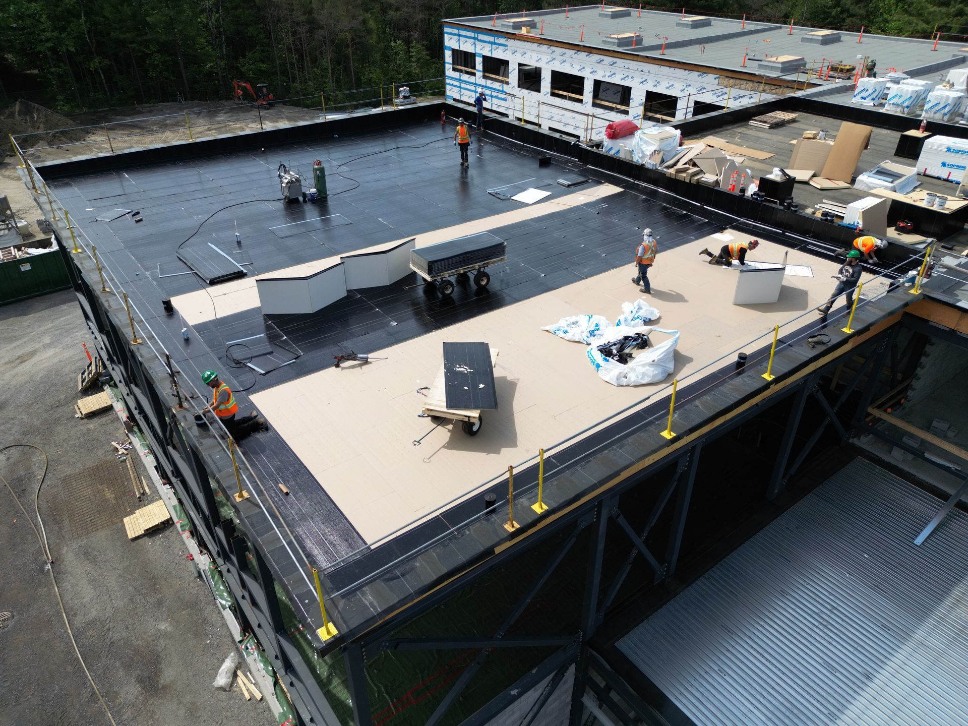 Rooftop worksite with workers, HVAC units, and construction materials on a flat black roof
